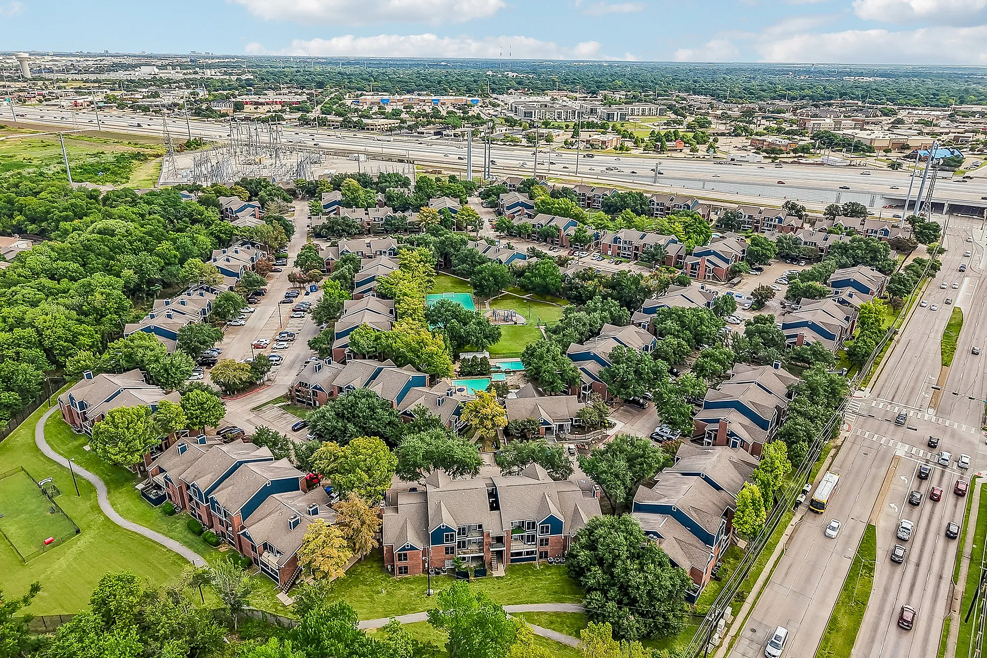 Scenic aerial view of a vibrant apartment complex with lush greenery, walking paths, and central pool area, located near major highway.