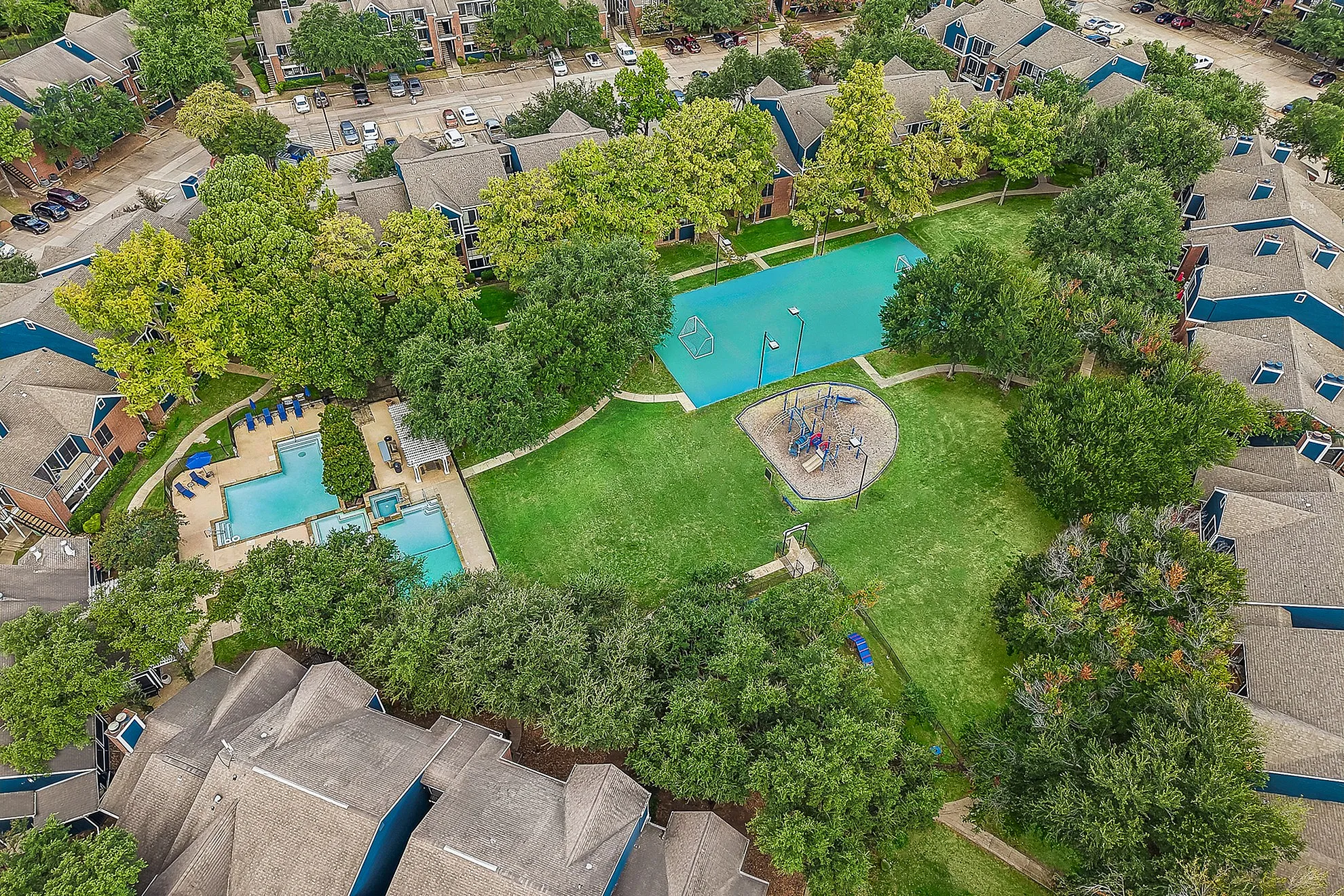 Aerial view of a vibrant apartment complex featuring lush greenery, a sparkling swimming pool, and a playful playground area, surrounded by charming residential buildings.