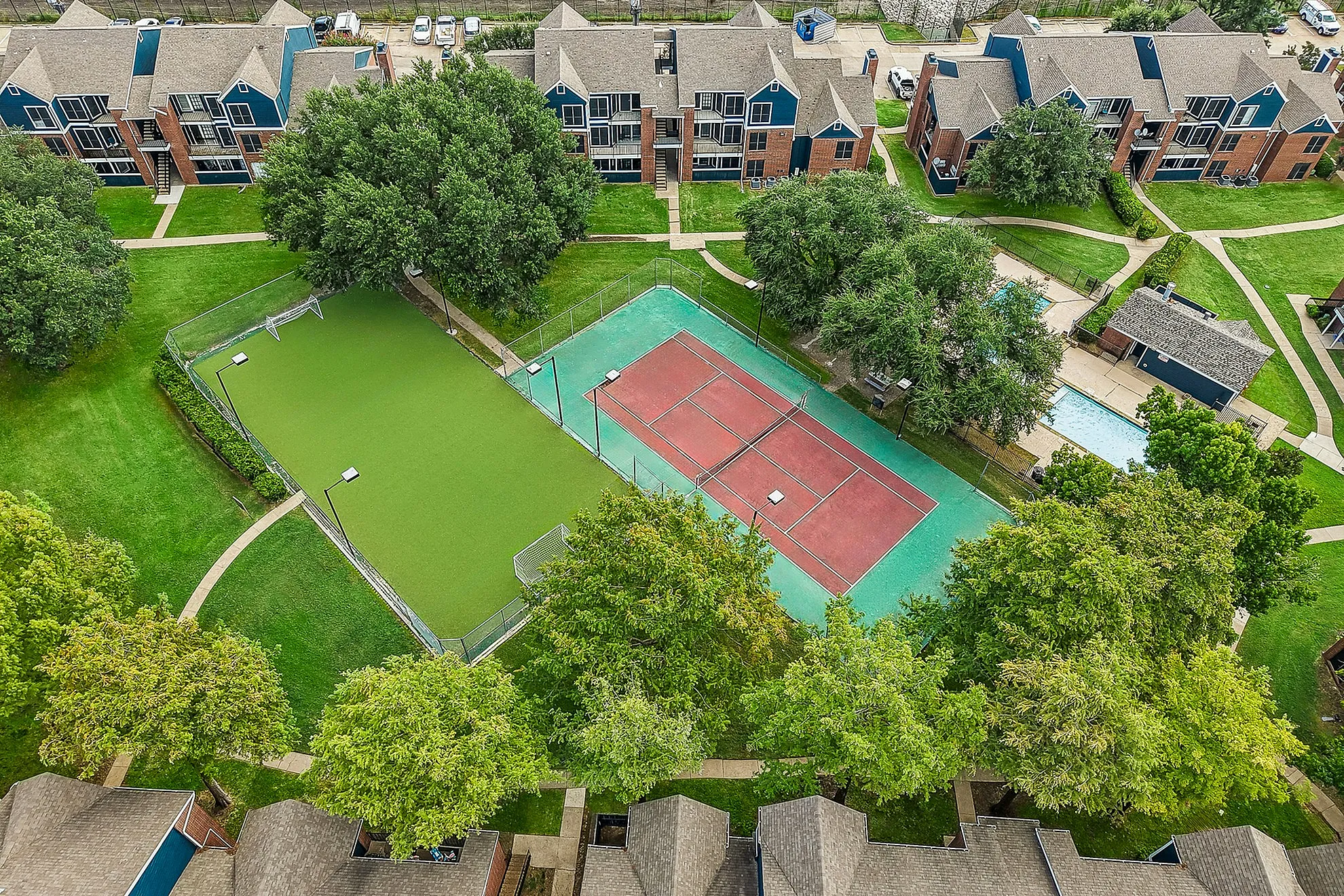 Scenic aerial view of modern apartment complex with lush greenery, a well-maintained tennis court, and a sparkling pool, offering vibrant community living.