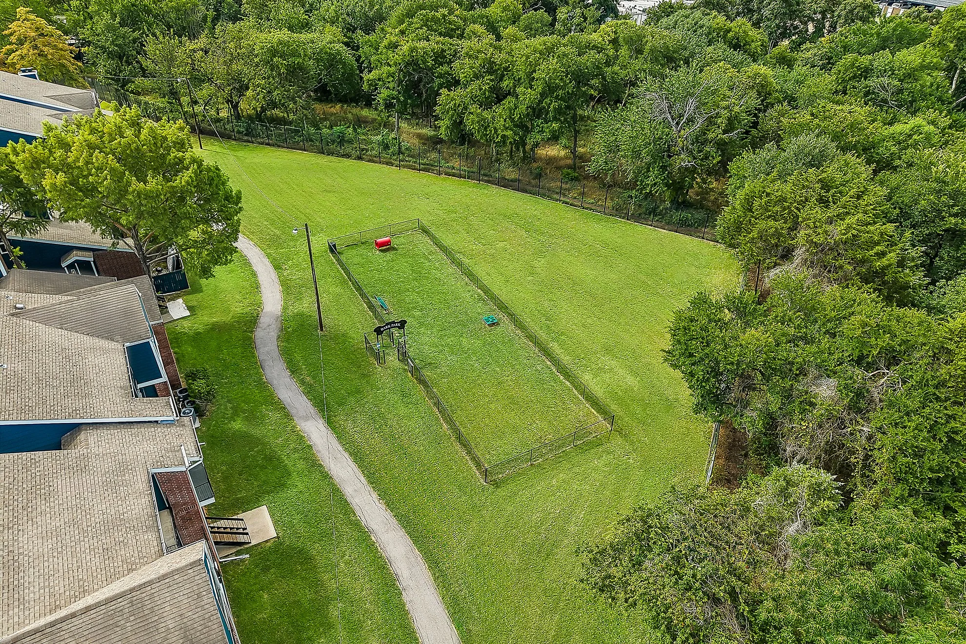 Serene aerial view of a lush green dog park surrounded by vibrant trees, adjacent to a charming apartment complex.