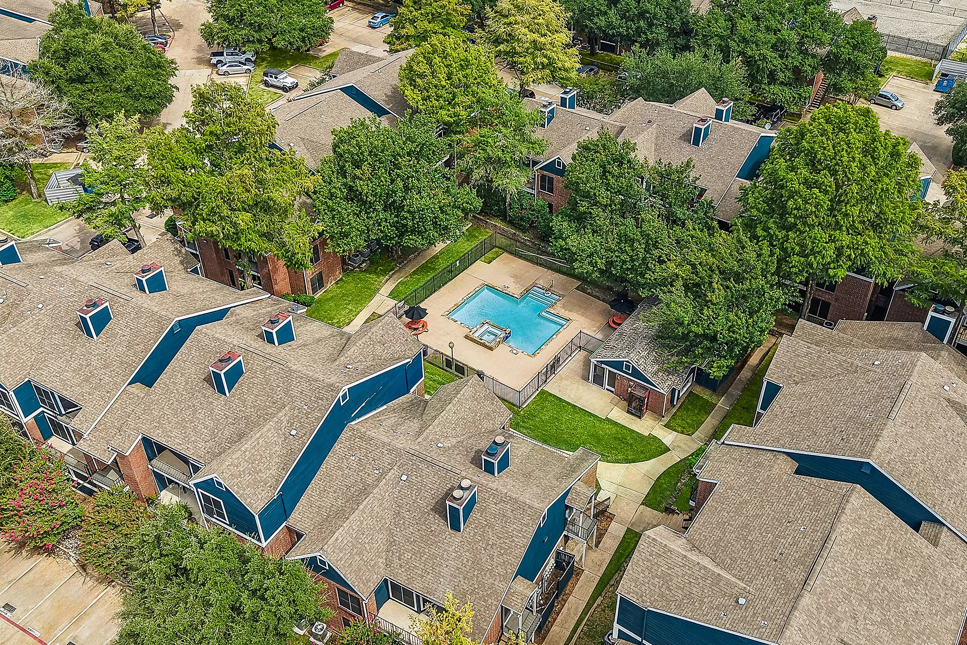 Aerial view of charming townhouse complex with navy-blue accents, nestled among lush greenery, featuring a central swimming pool for relaxation.