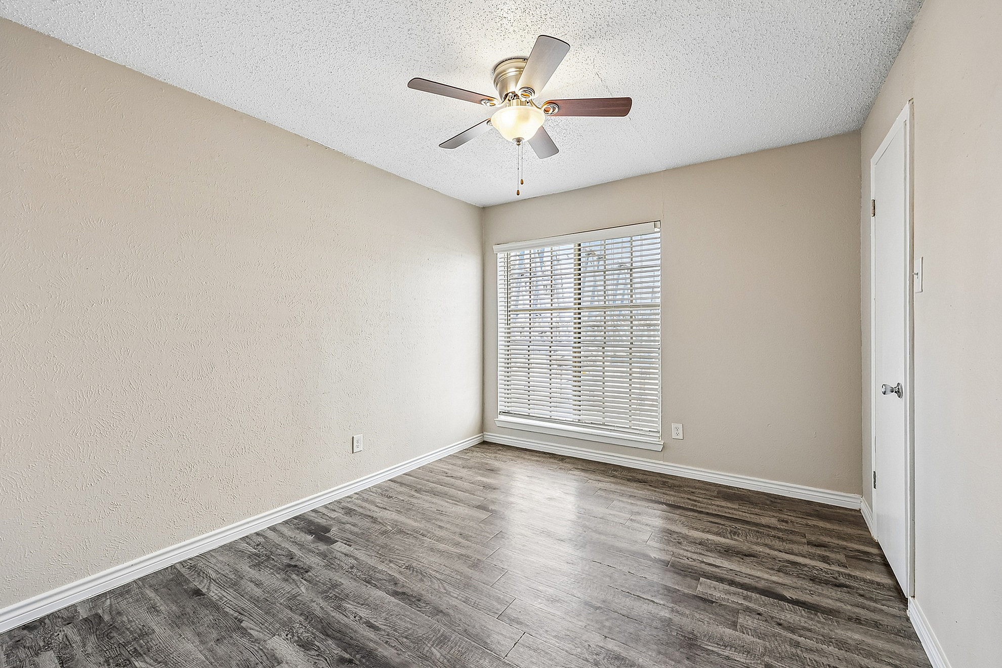 Bright bedroom with elegant wood flooring, a large window with blinds, and a stylish ceiling fan, offering a comfortable and airy living space.