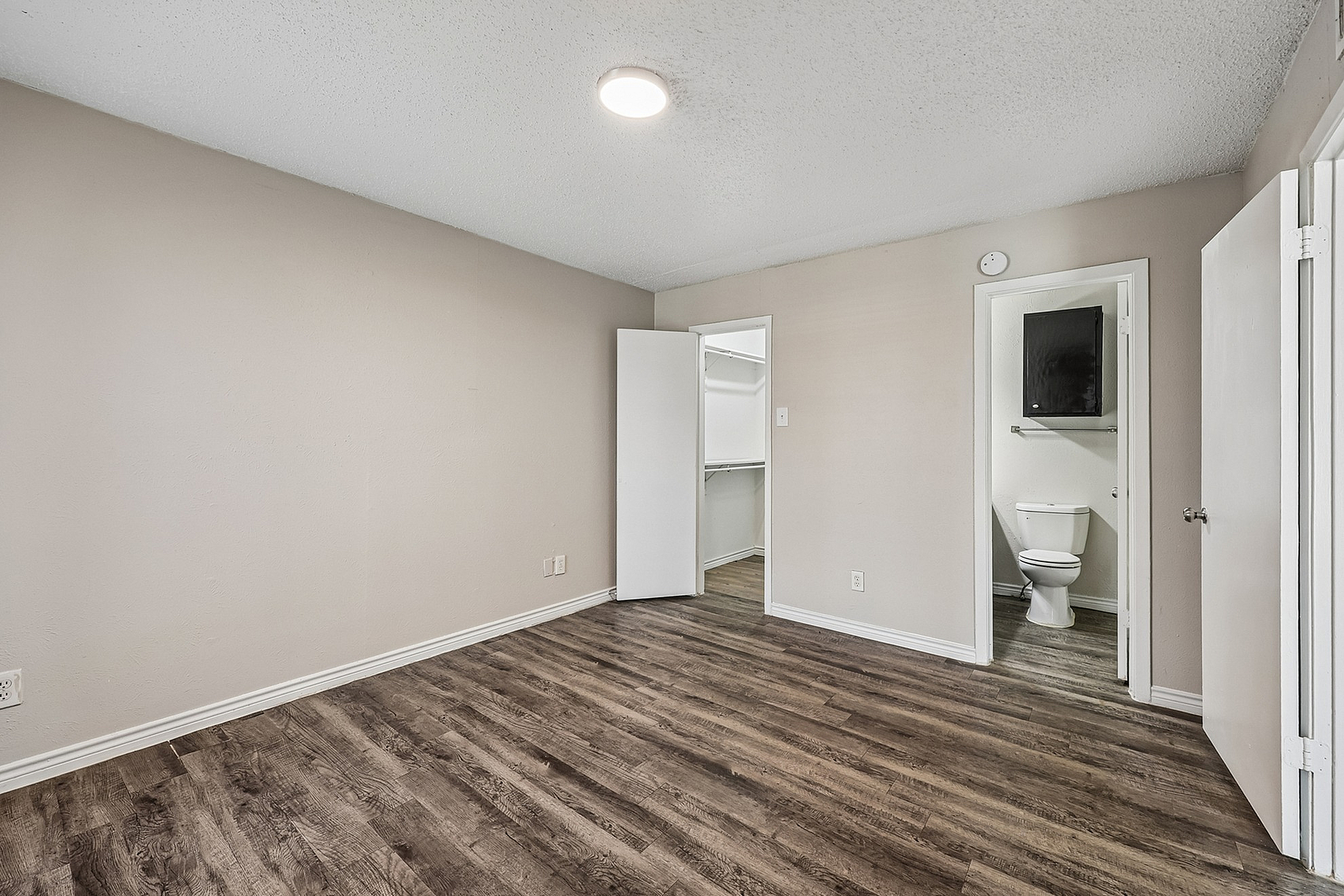 Spacious room with elegant wood flooring, bright lighting, and convenient access to a walk-in closet and bathroom.