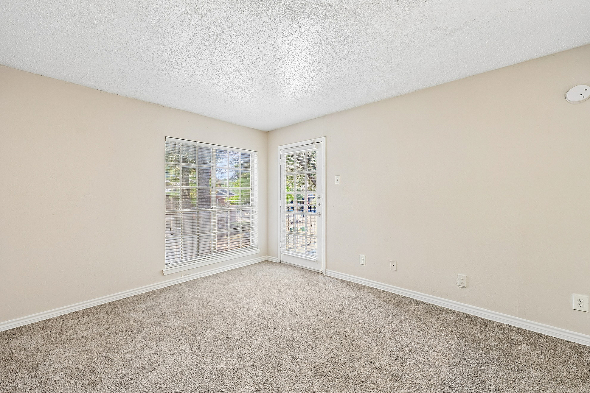 Bright and airy living room with plush carpeting, featuring large windows and a glass door opening to a scenic outdoor view, perfect for relaxation.