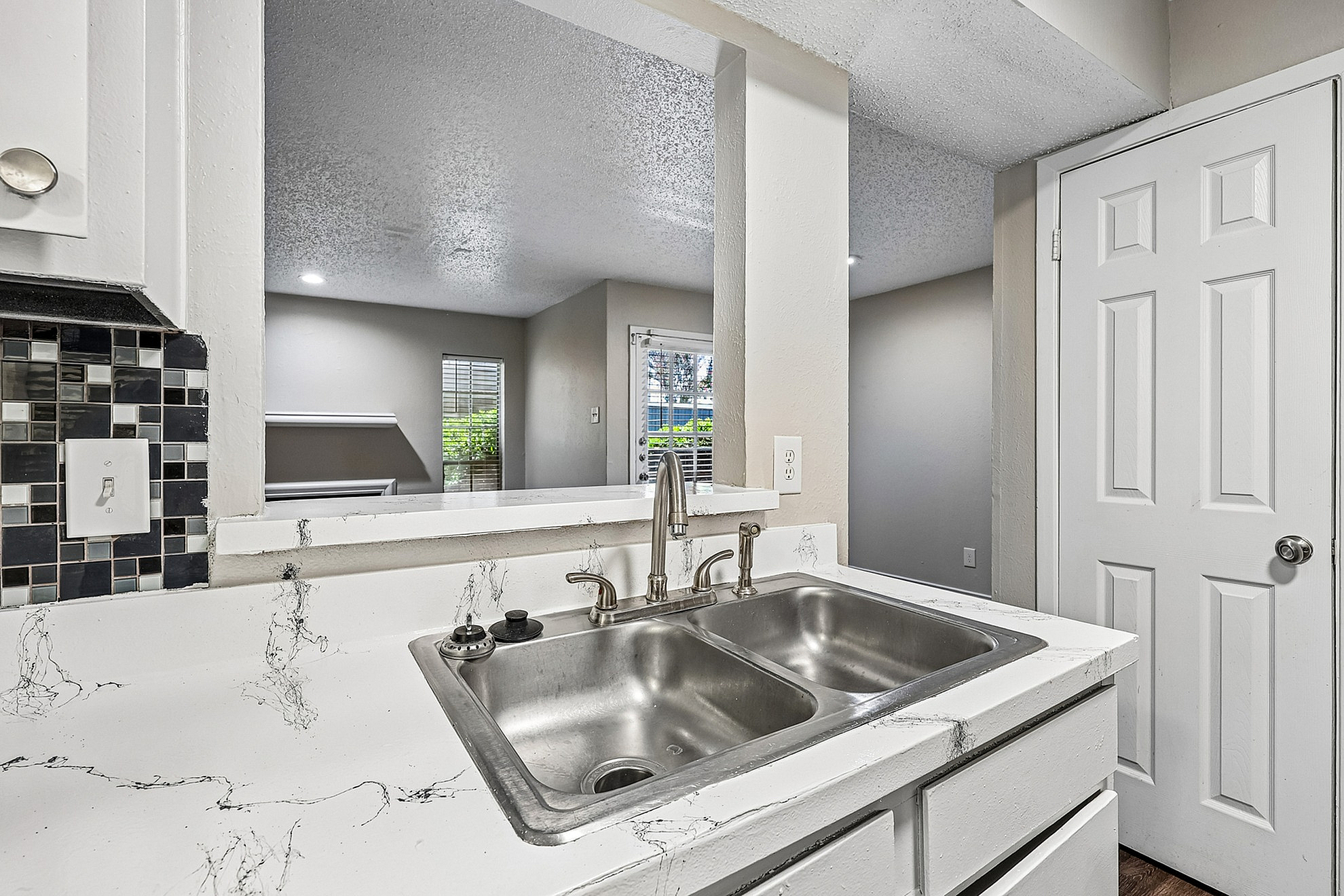 Modern kitchen with a sleek dual-basin sink, elegant marble countertops, and stylish backsplash, offering a glimpse into a sunlit living area.