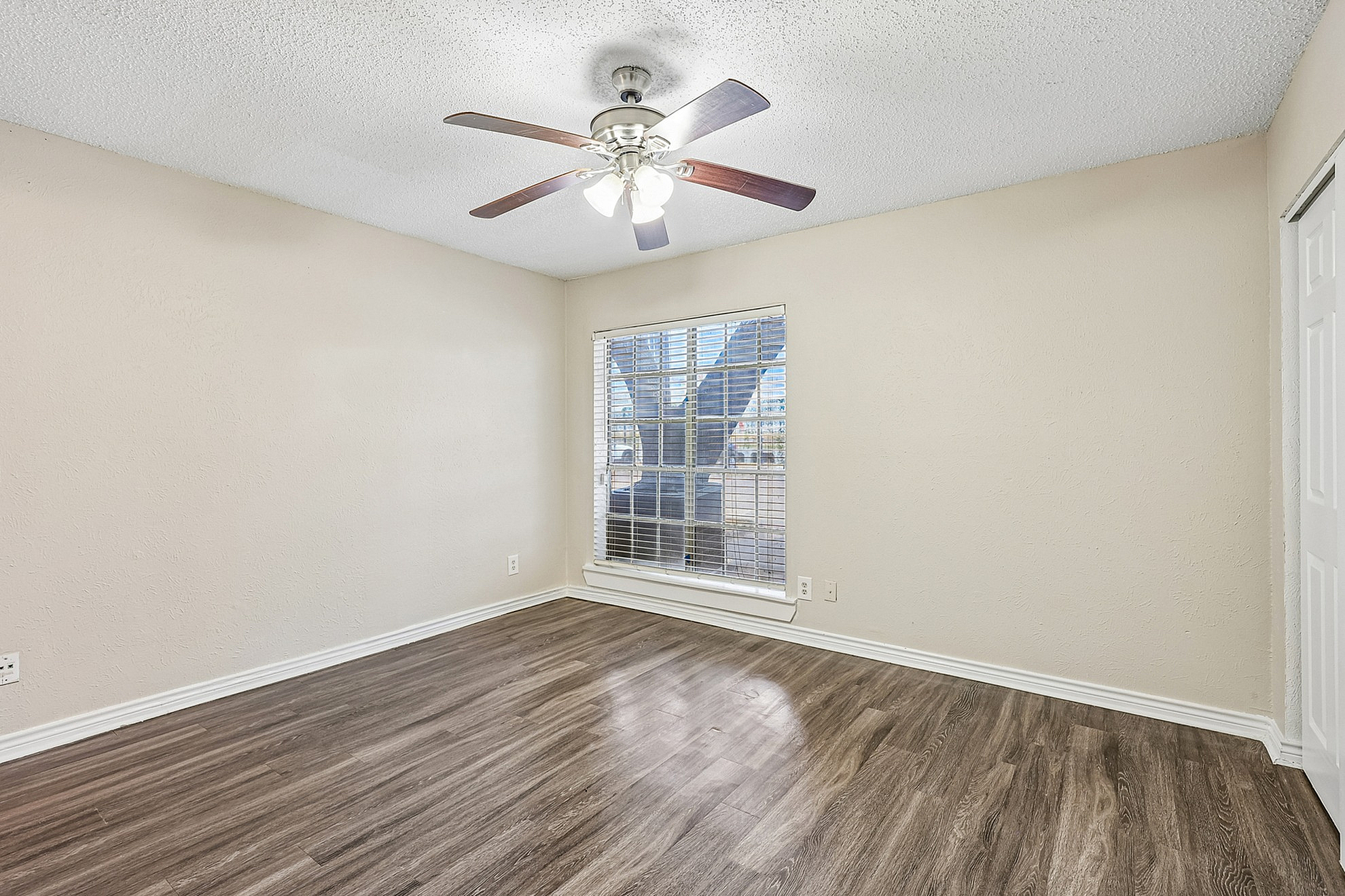 Charming vacant room with stylish wooden flooring, neutral walls, a ceiling fan, and a bright window view.