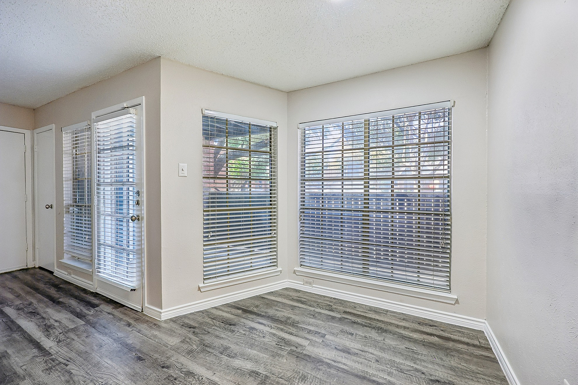 Bright corner room with elegant wood flooring and large windows with blinds, offering ample natural light and a cozy atmosphere.