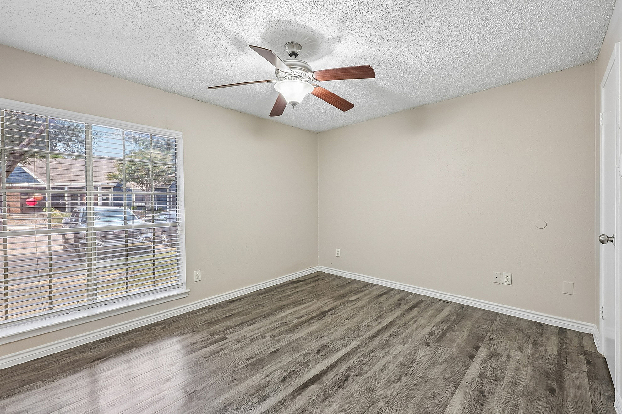 Bright apartment room featuring elegant wooden flooring, a ceiling fan, and a large window offering a view of the peaceful neighborhood.