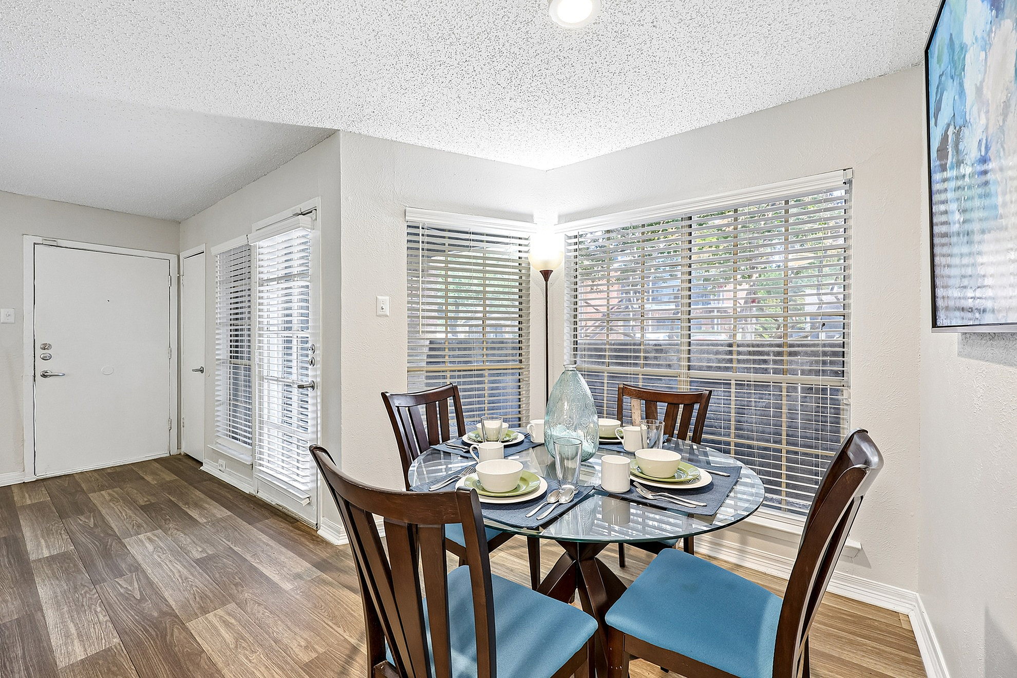 Charming dining area with a round glass table set for four, featuring elegant wood chairs and natural light streaming through large windows with blinds.