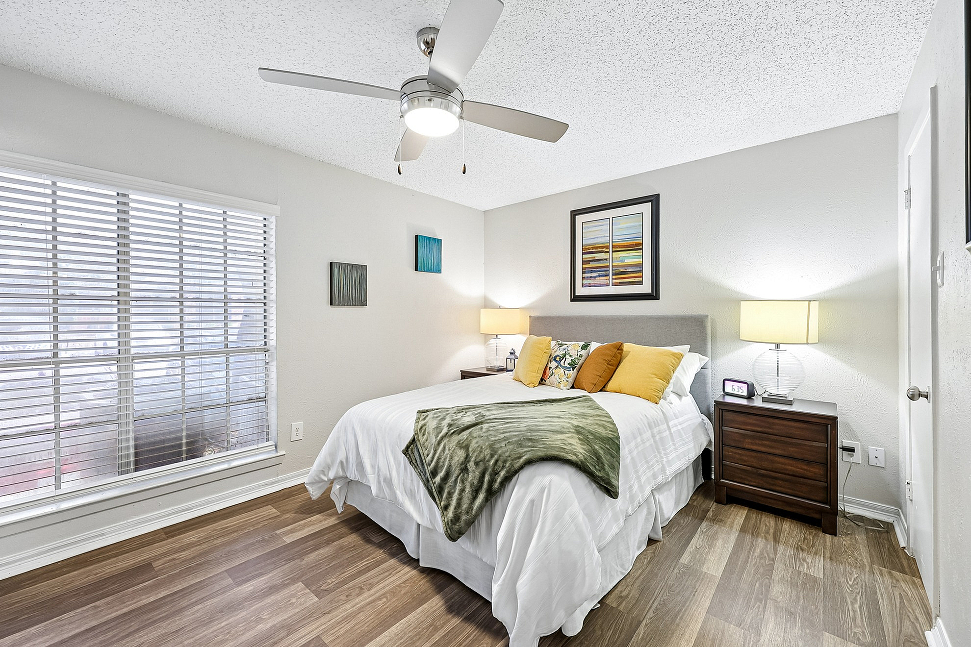 Inviting bedroom with elegant wood flooring, modern ceiling fan, and tasteful decor, enhanced by ample natural light through large windows.