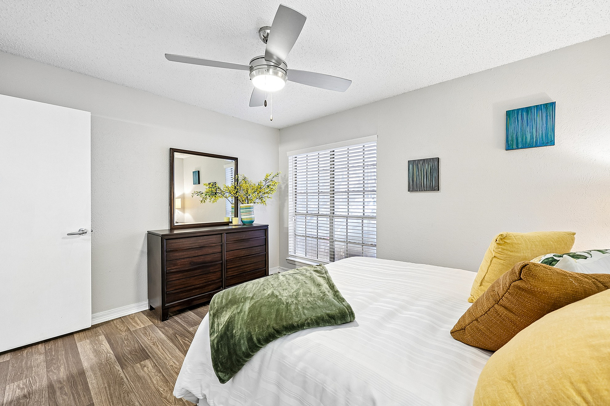 Bright and inviting bedroom with modern decor, featuring a plush bed, stylish ceiling fan, and chic wooden dresser against a sunny window backdrop.