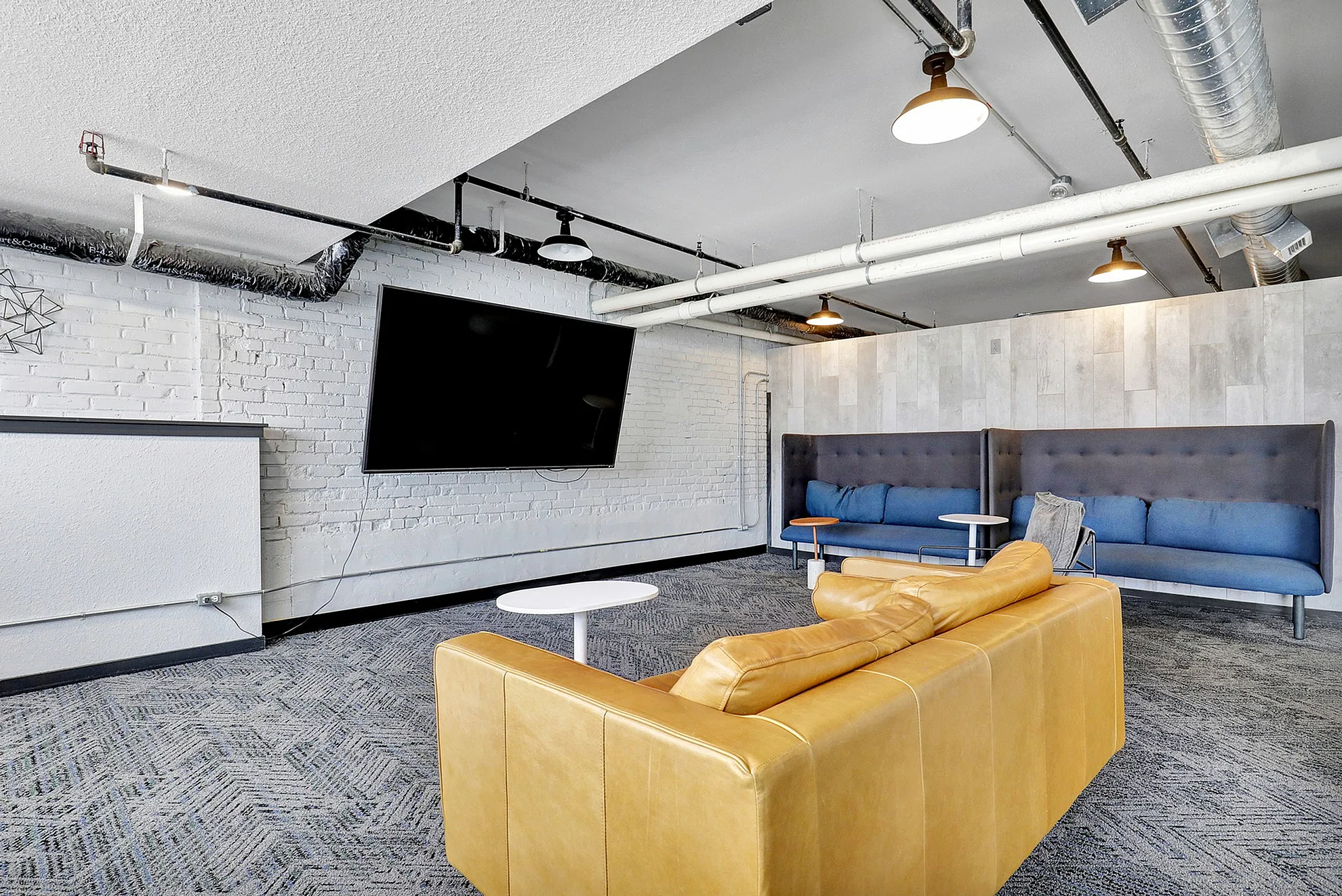 Modern industrial-style lounge with a large flat-screen TV, cozy yellow sofa, and sleek blue seating against a chic brick wall.