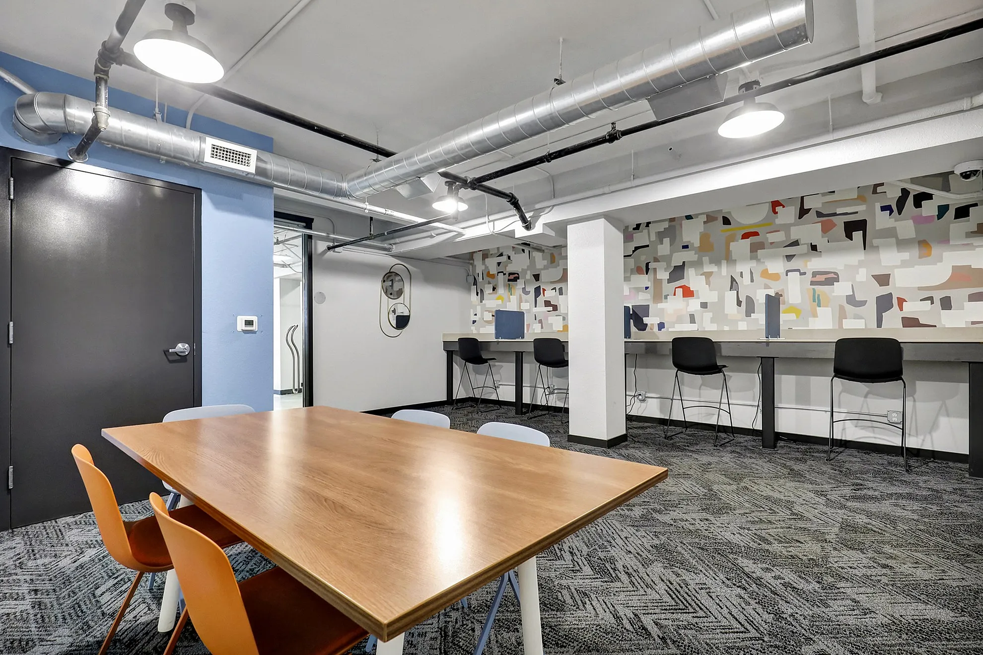 Modern coworking space with chic decor, featuring a large wooden table, colorful chairs, and a vibrant abstract mural.
