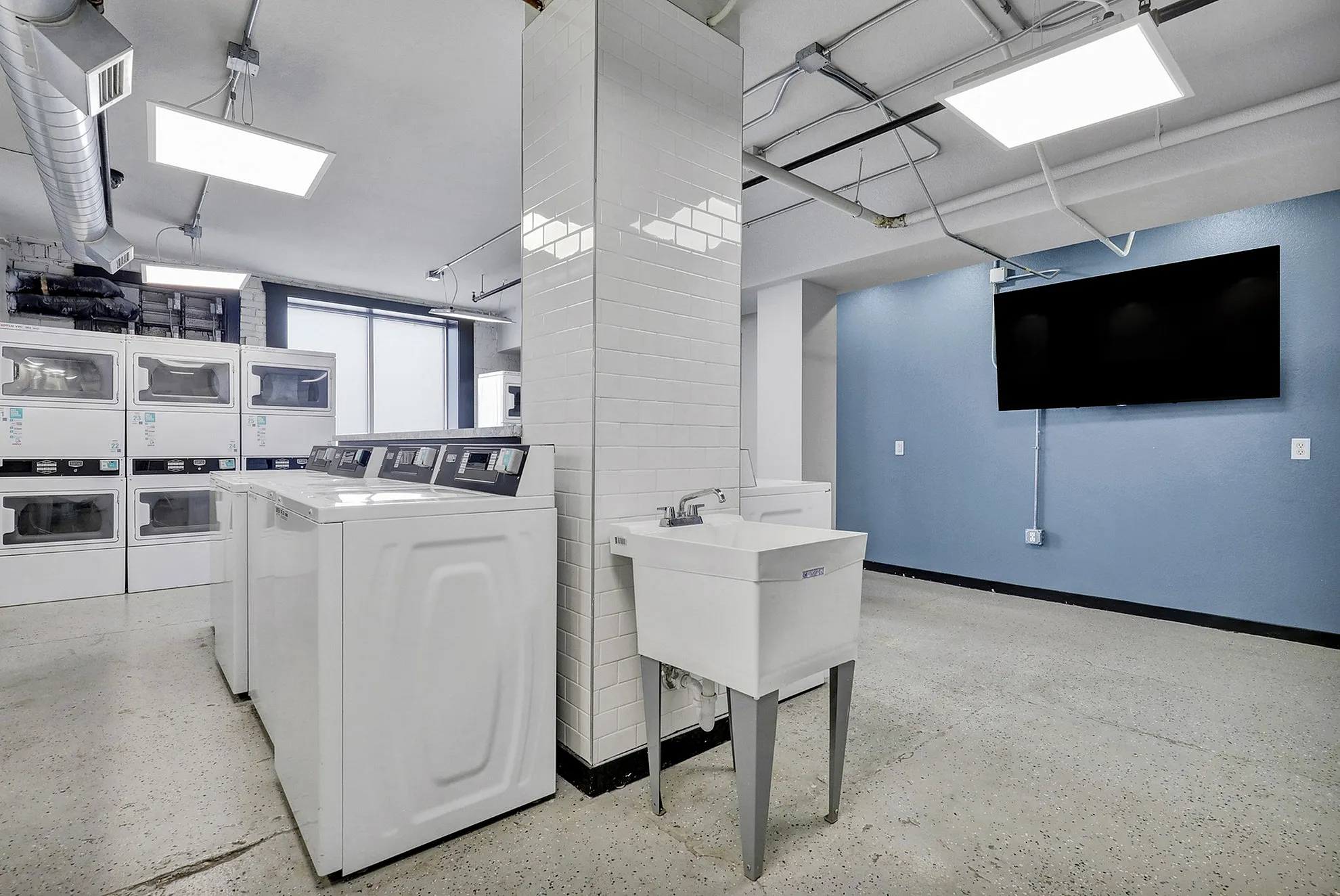 Modern laundry room with multiple washers and dryers, featuring a sleek utility sink and a flat-screen TV against a stylish blue accent wall.