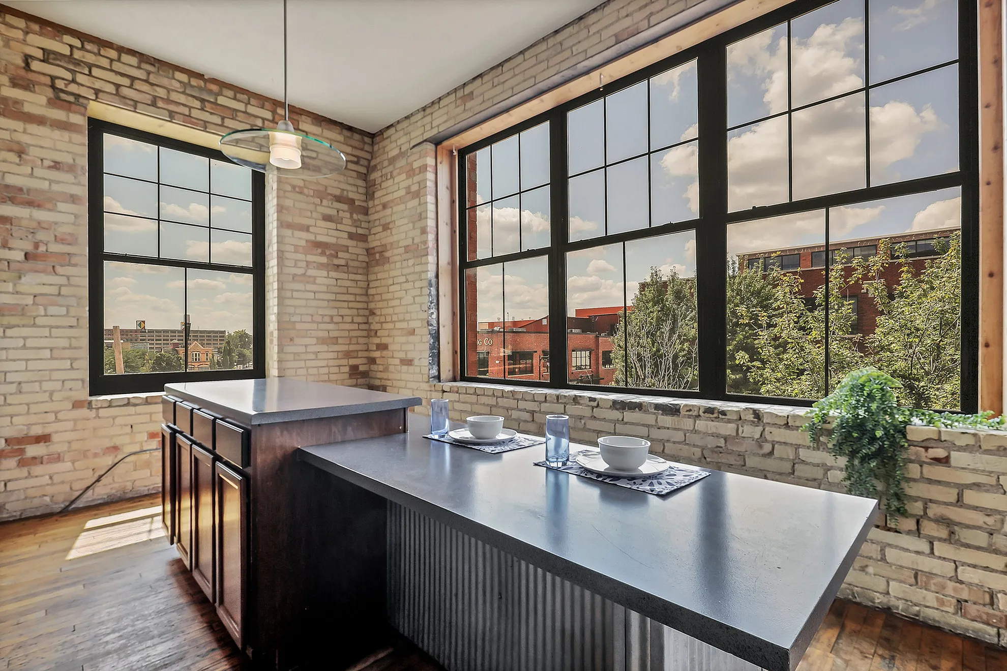 Charming loft kitchen with exposed brick walls, large windows offering city views, and a sleek breakfast bar with modern place settings.