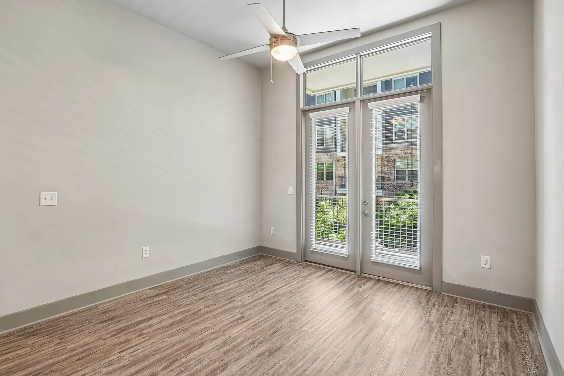 Bright, empty apartment room with wooden floors, large windows, and a ceiling fan, offering modern and sleek design.