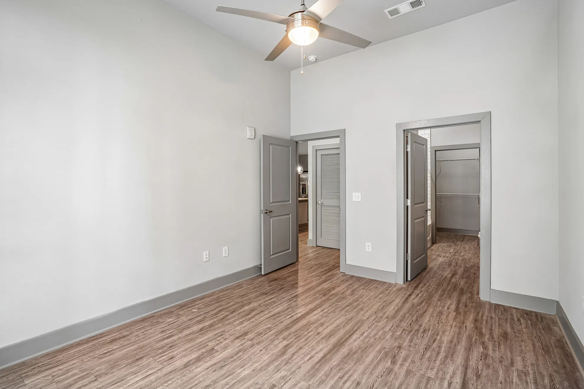 Modern, empty room with elegant wood flooring and a ceiling fan, leading to a spacious walk-in closet and bathroom area.