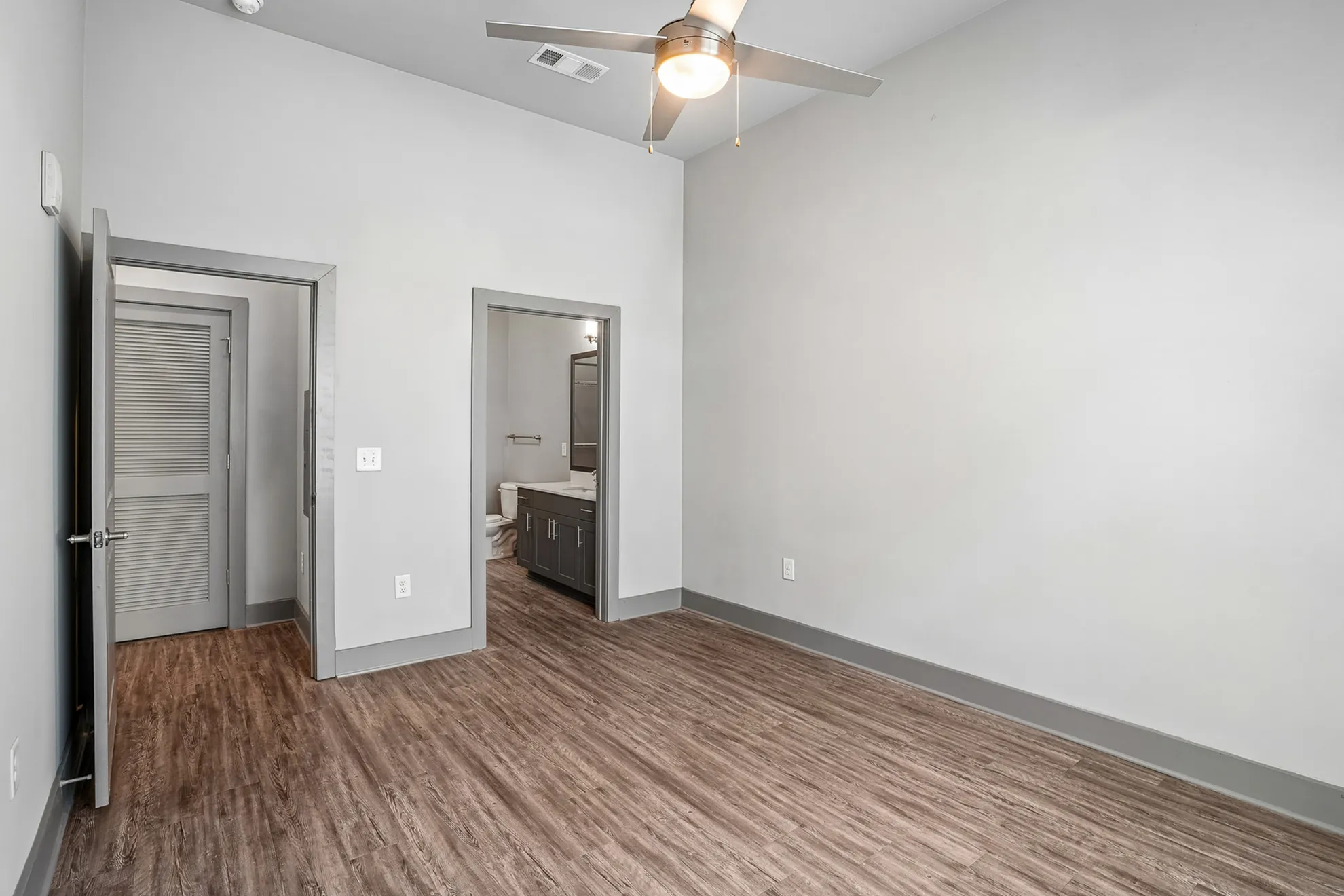 Modern bedroom featuring sleek hardwood floors, neutral gray walls, and an en-suite bathroom, illuminated by a stylish ceiling fan light.