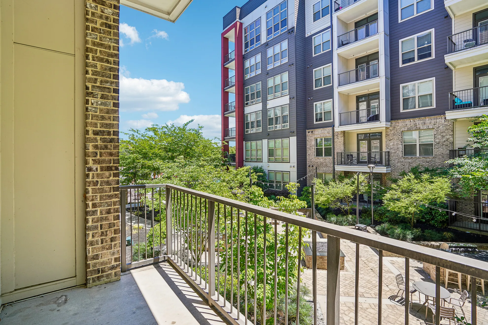 Charming apartment balcony view overlooking lush gardens and modern building fa&ccedil;ade under a bright blue sky.