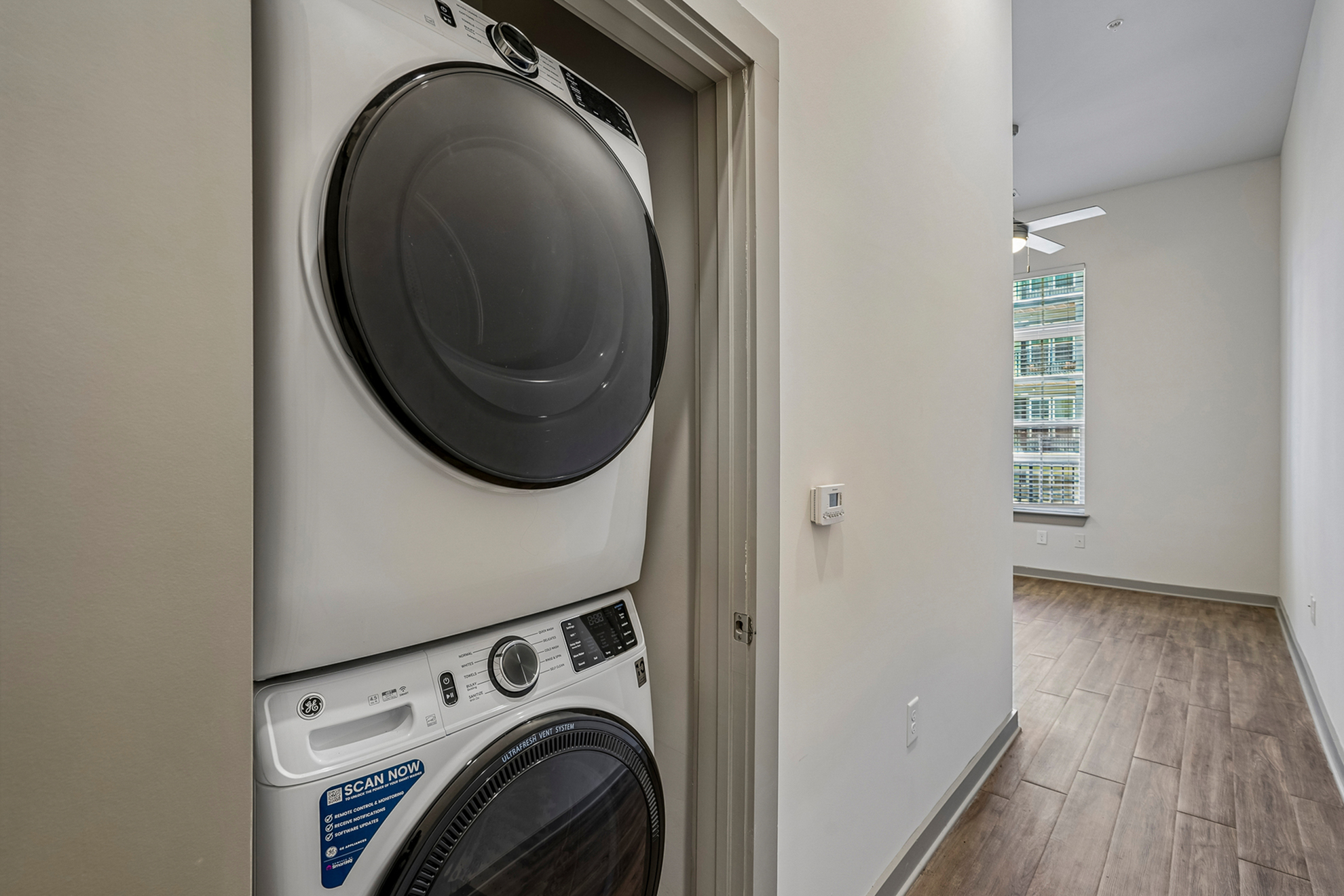 Contemporary apartment hallway featuring an in-unit stacked washer and dryer next to a bright room with elegant wood flooring and large windows.