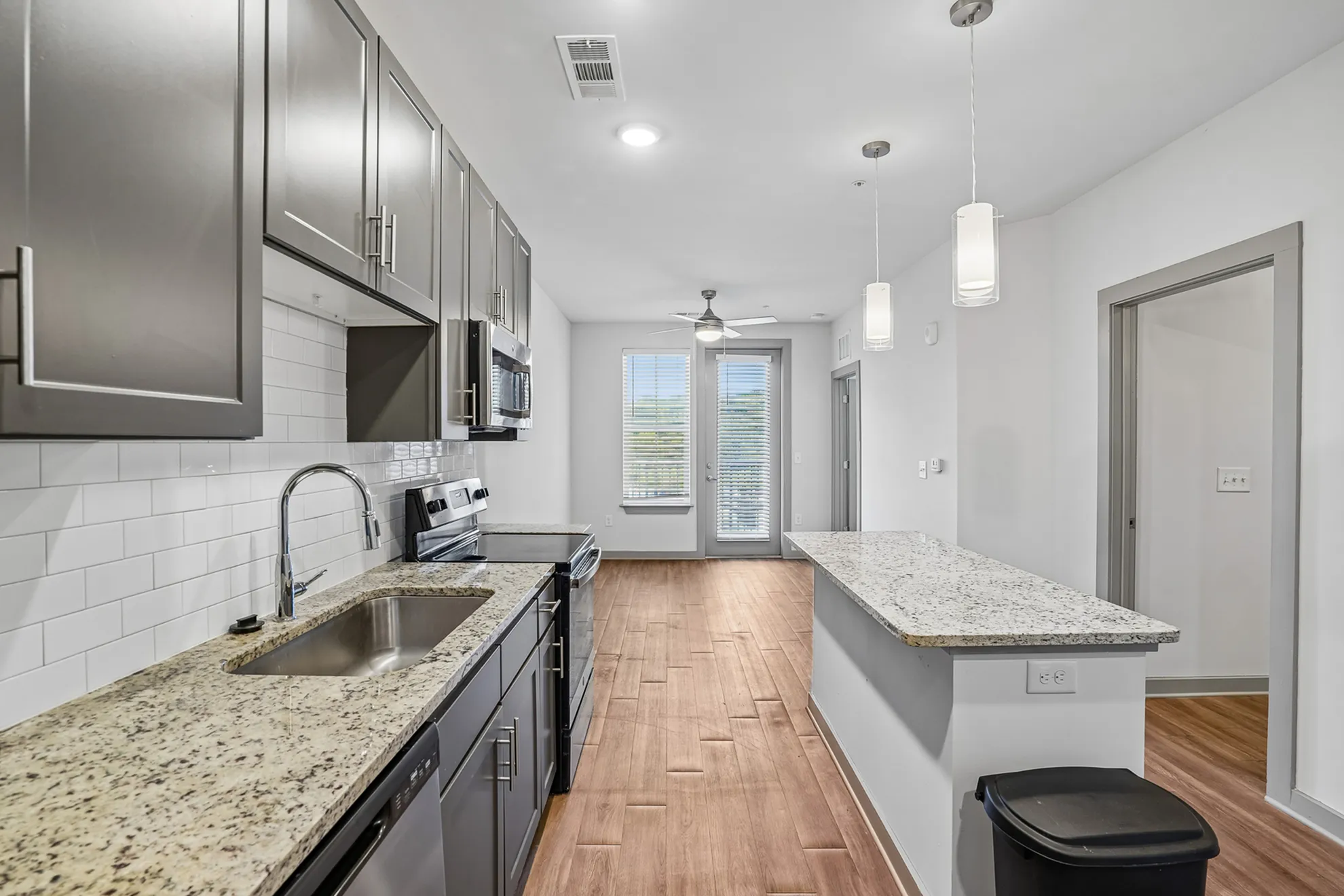 Modern kitchen with sleek cabinetry, granite countertops, and stainless steel appliances, leading to a bright living area with natural light from large windows.