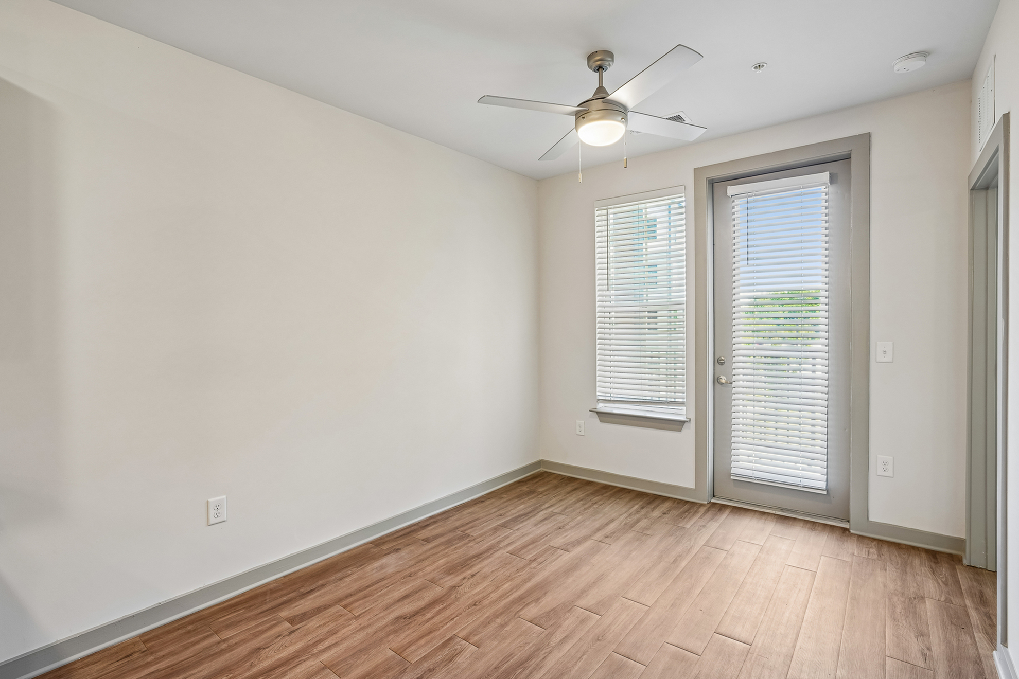 Bright and modern room featuring sleek wooden floors, large windows with blinds, and a contemporary ceiling fan.