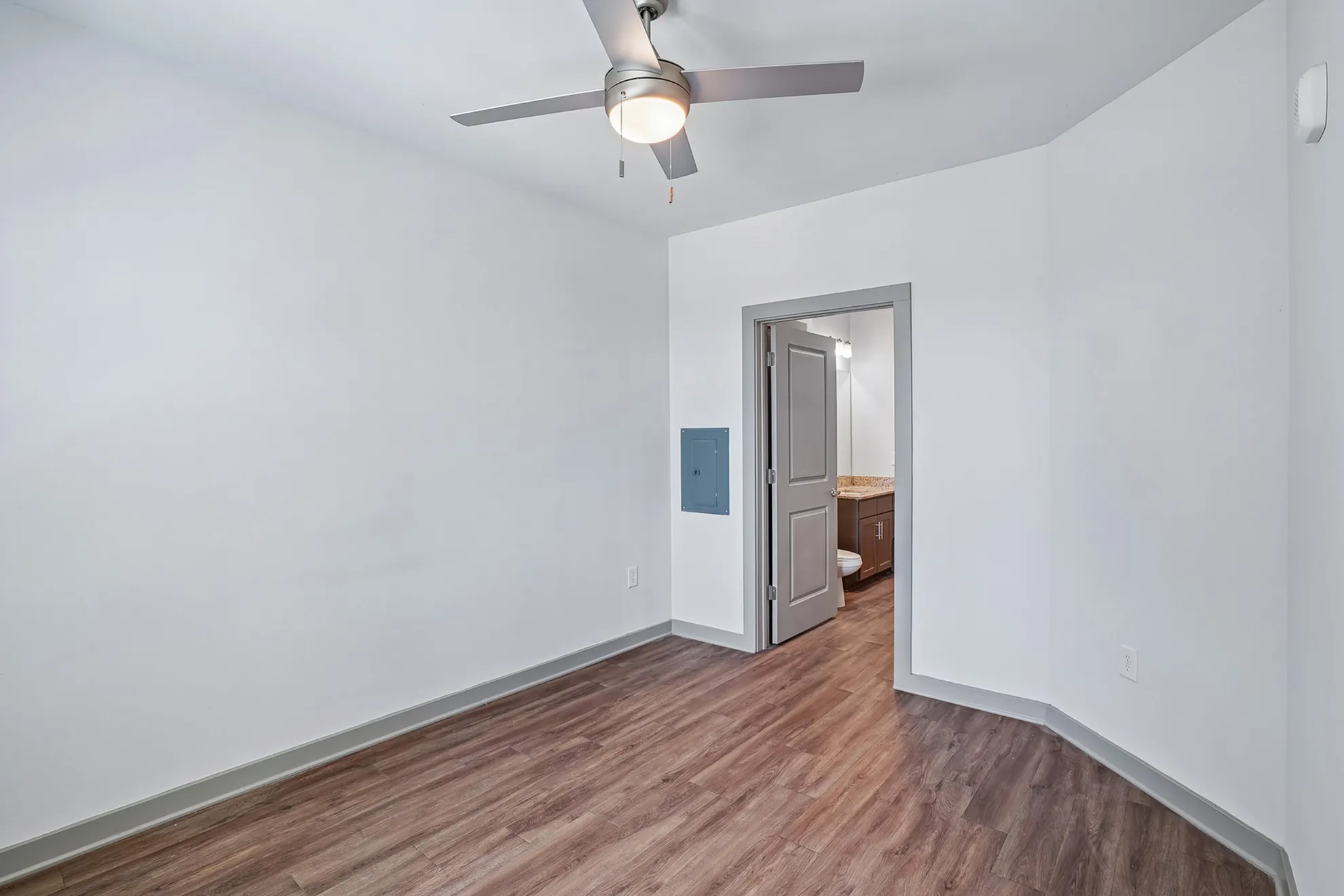 Bright and inviting room with modern wood flooring and a sleek ceiling fan, leading to a well-lit bathroom with contemporary fixtures.