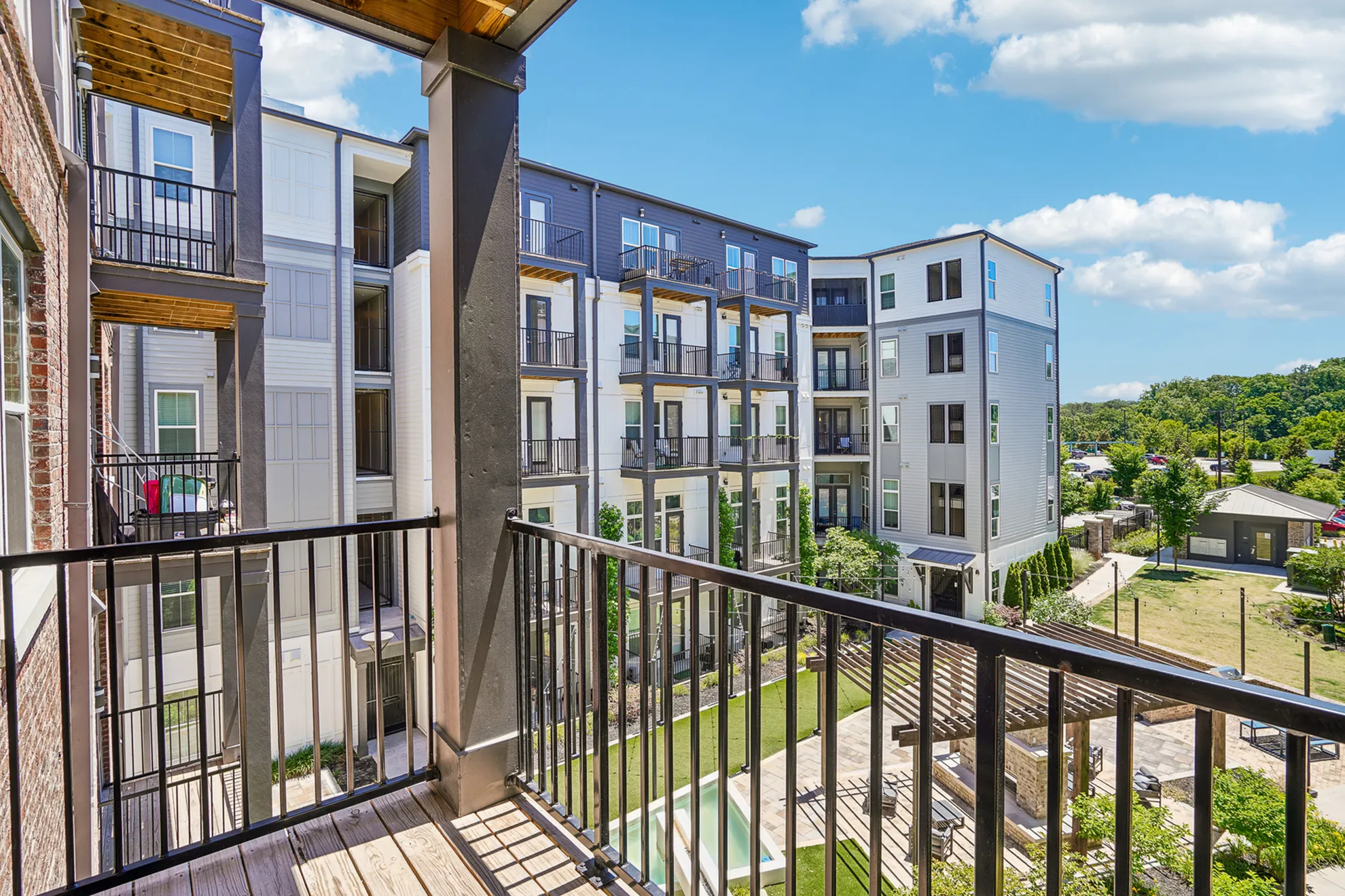 Modern apartment balcony with a stunning view of a landscaped courtyard, surrounded by contemporary multi-story buildings under a bright blue sky.