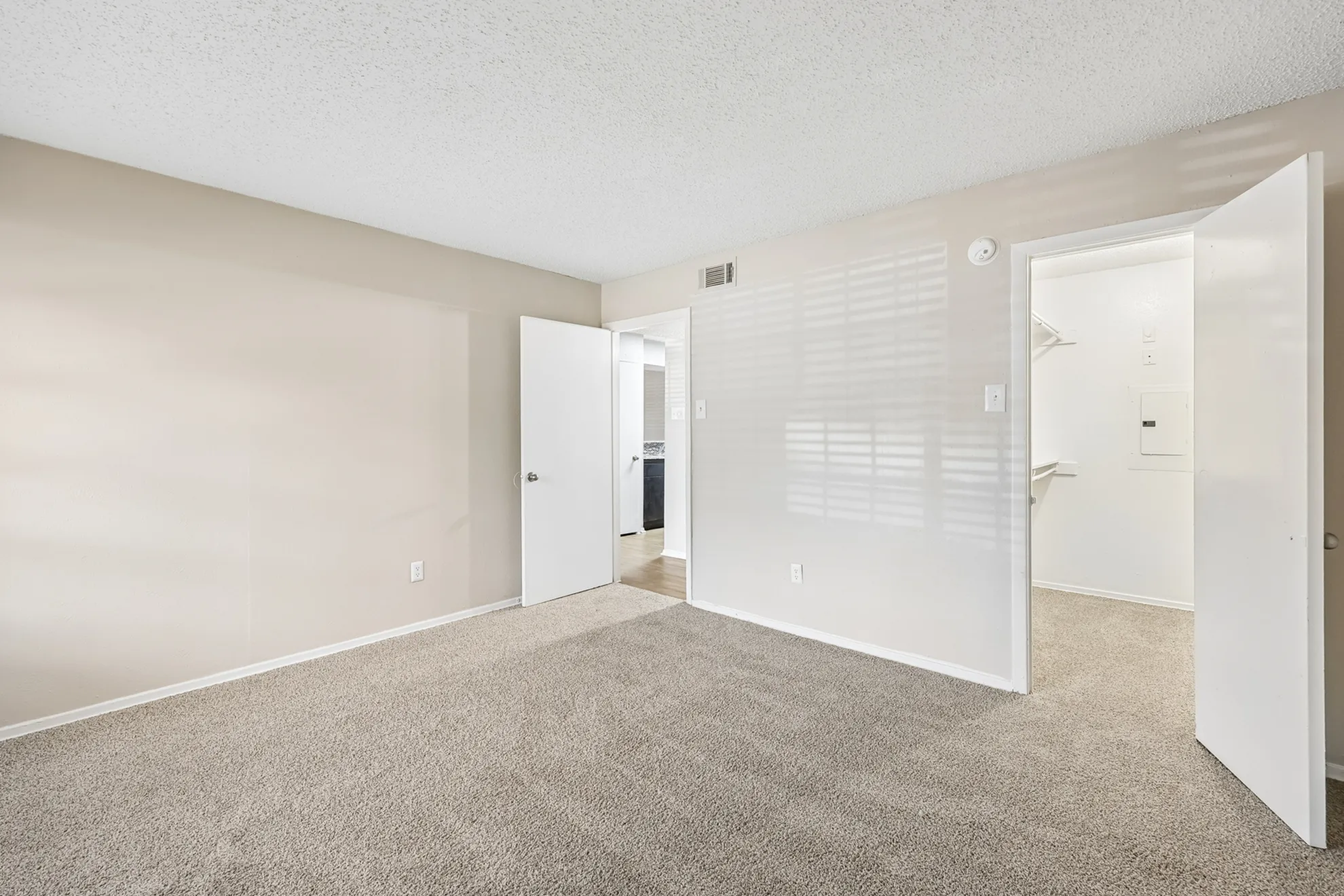 Bright and spacious bedroom with neutral carpeting and ample natural light, featuring an open walk-in closet and a view into an adjacent room.