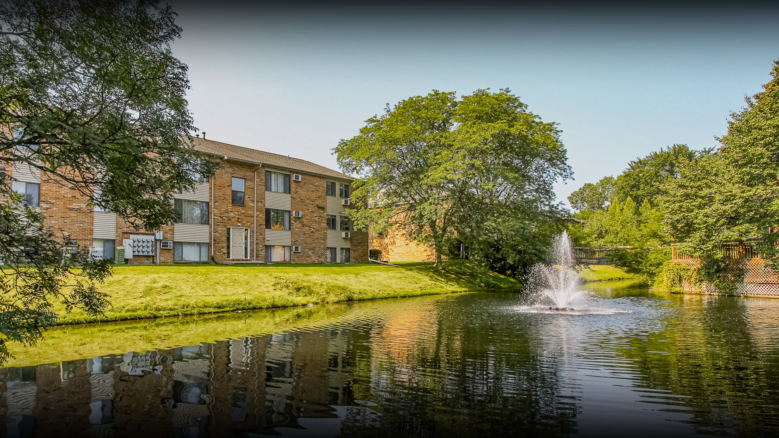 Serene apartment complex with lush greenery, a shimmering pond, and a beautiful fountain reflecting peaceful living.