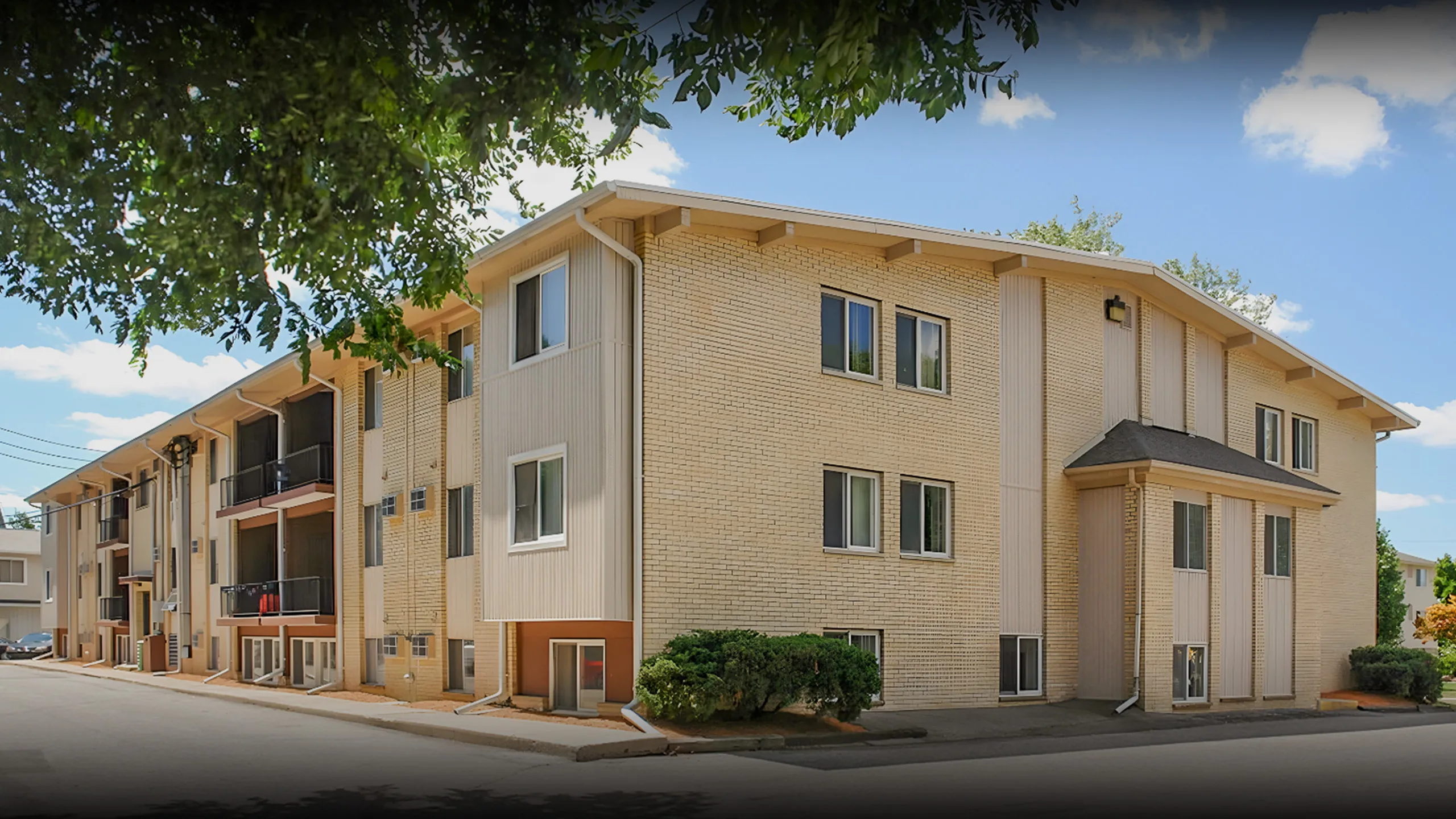 Charming multi-story apartment building with brick exterior, nestled in a serene neighborhood with lush greenery and expansive blue sky.