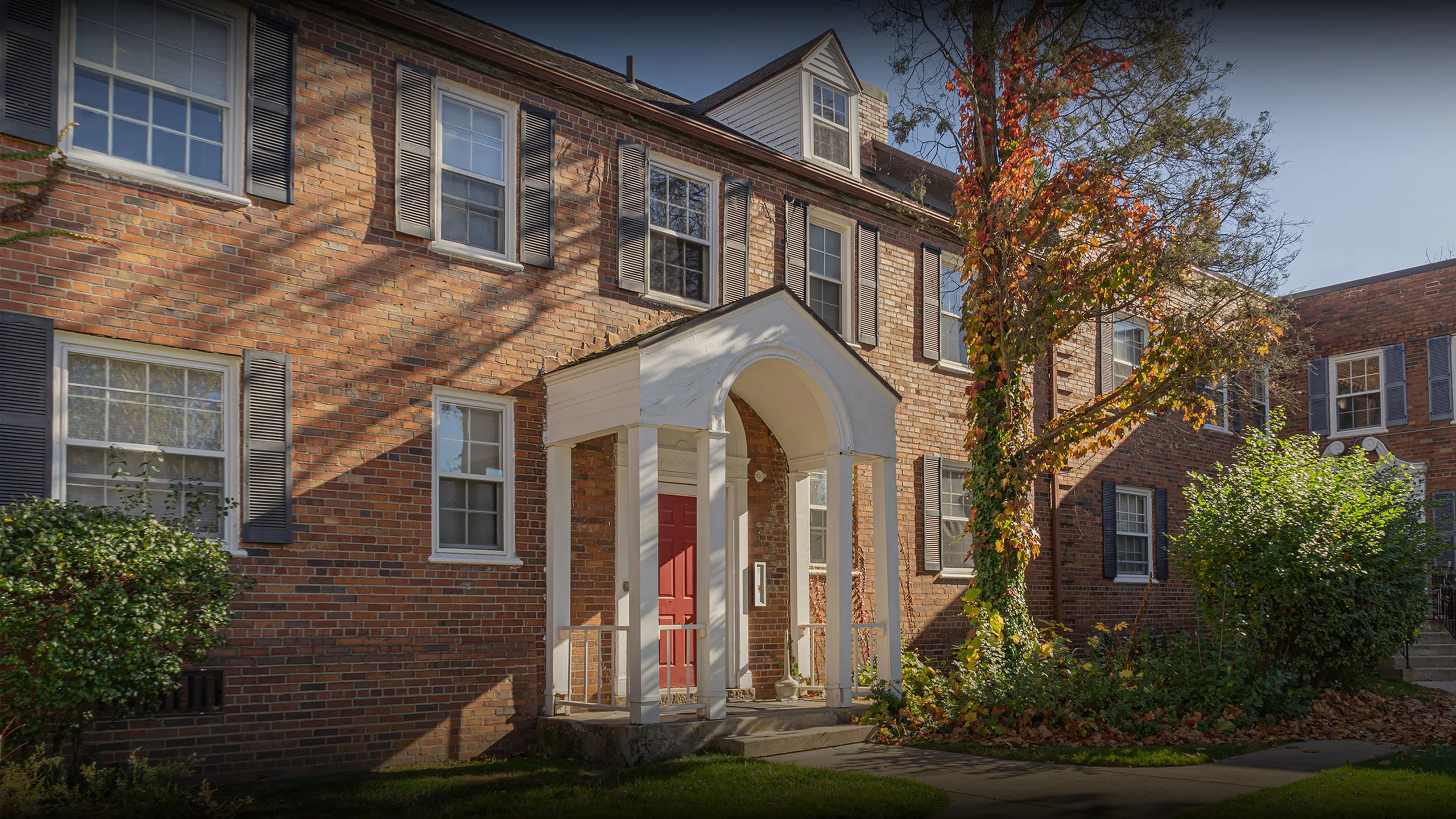 Charming brick apartment building with classic white columns and a vibrant red door, framed by lush greenery and sunlit exterior.