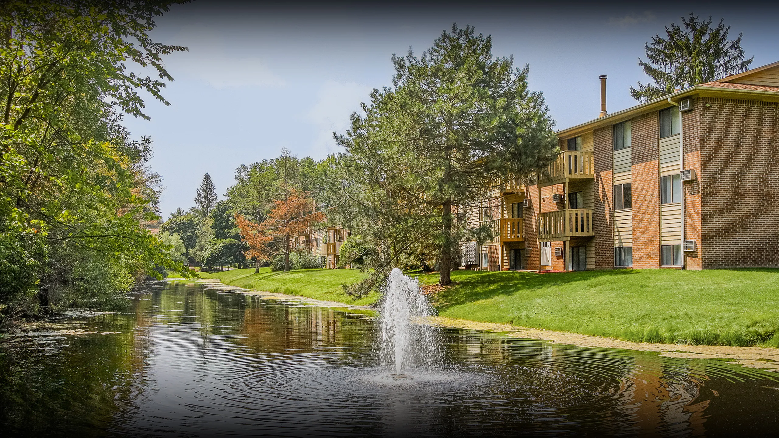 Charming apartment complex with brick exterior, nestled beside a serene pond and lush greenery, featuring a delightful fountain.