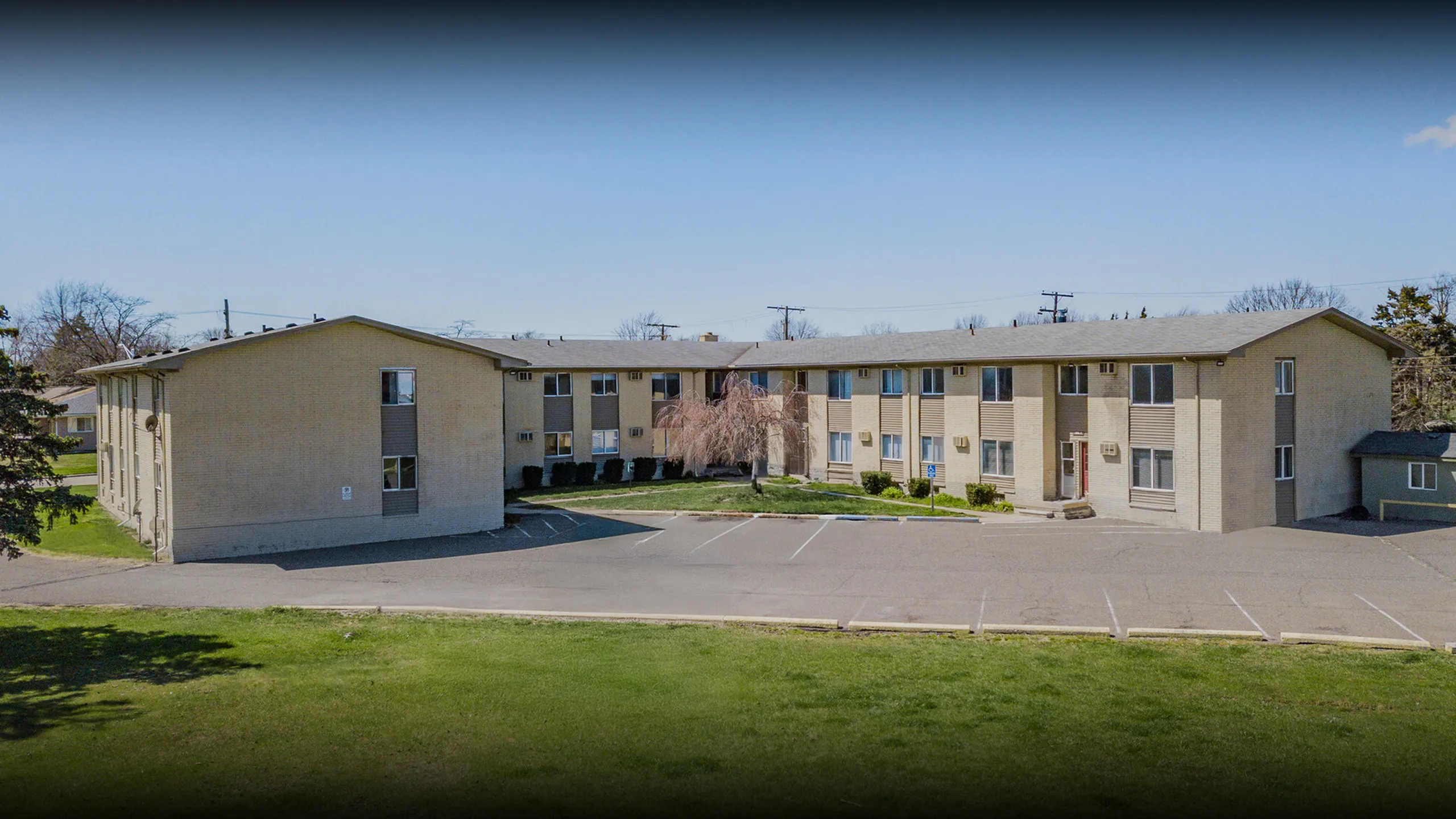 Charming two-story apartment building with ample parking, surrounded by lush greenery and clear blue skies.