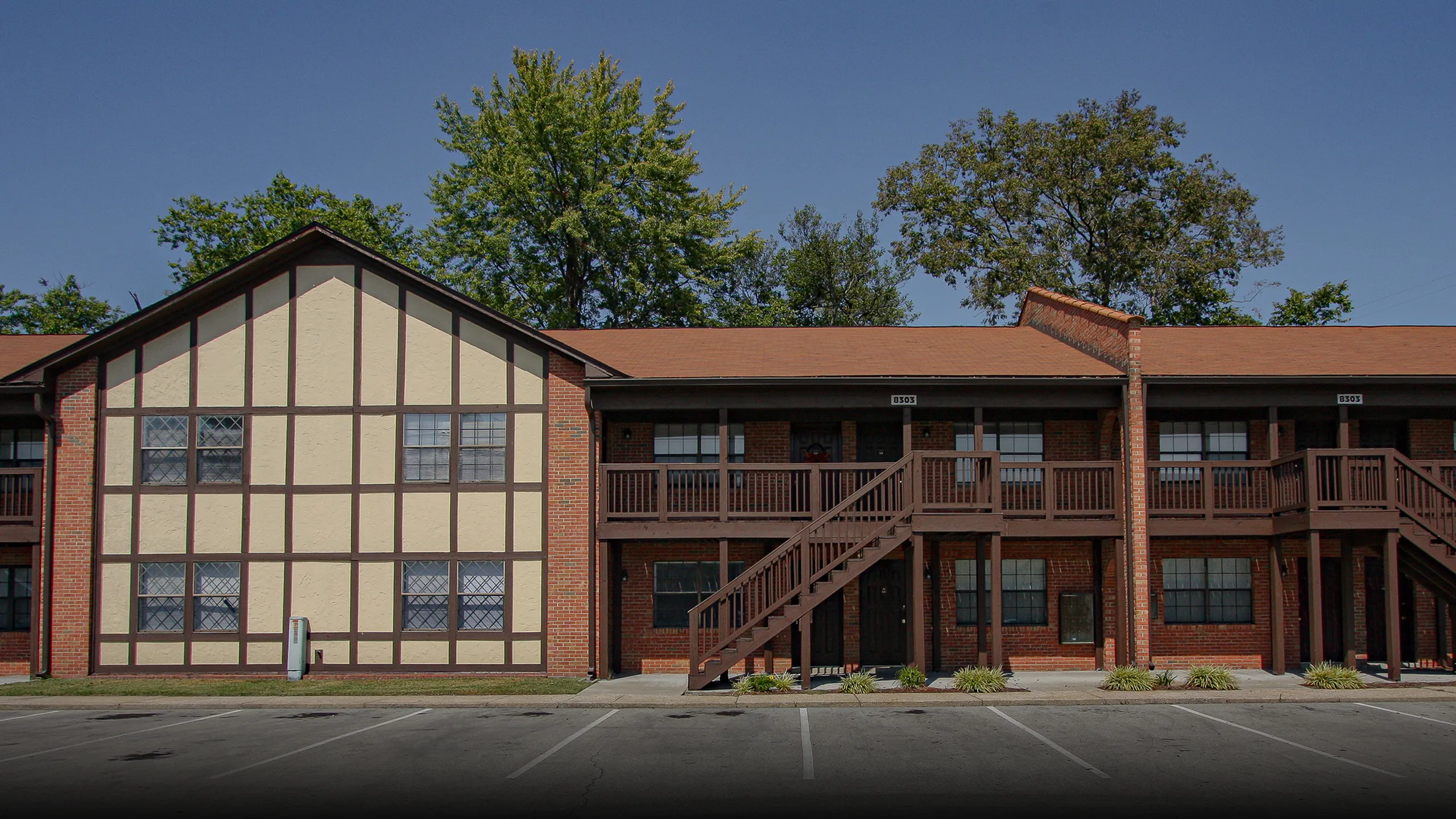 Charming two-story apartment building with rustic wooden balconies and Tudor-style accents, surrounded by lush greenery.
