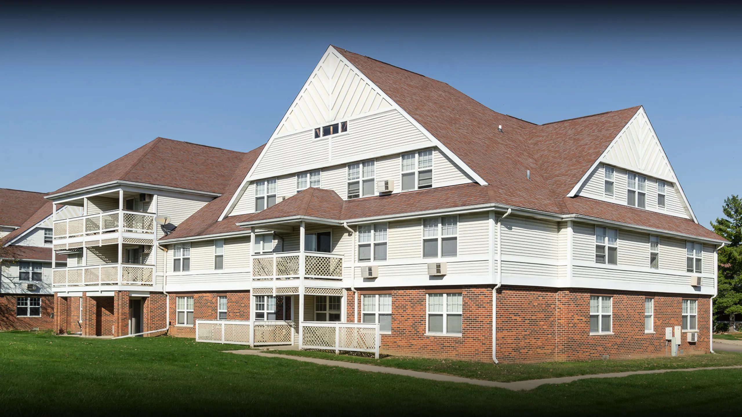 Charming three-story apartment complex with classic brick and siding design, featuring multiple balconies and lush green surroundings.