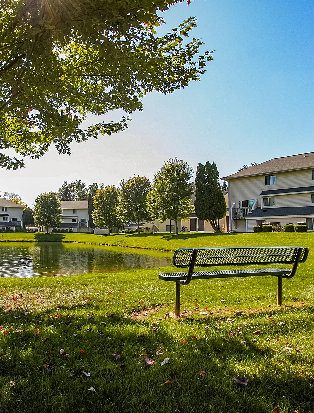 Tranquil apartment community with scenic pond view, lush greenery, and inviting bench under a sunlit tree.
