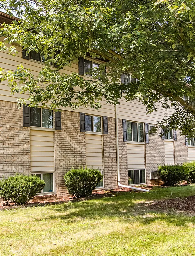 Charming brick apartment building surrounded by lush green landscaping and mature trees, offering serene suburban living.