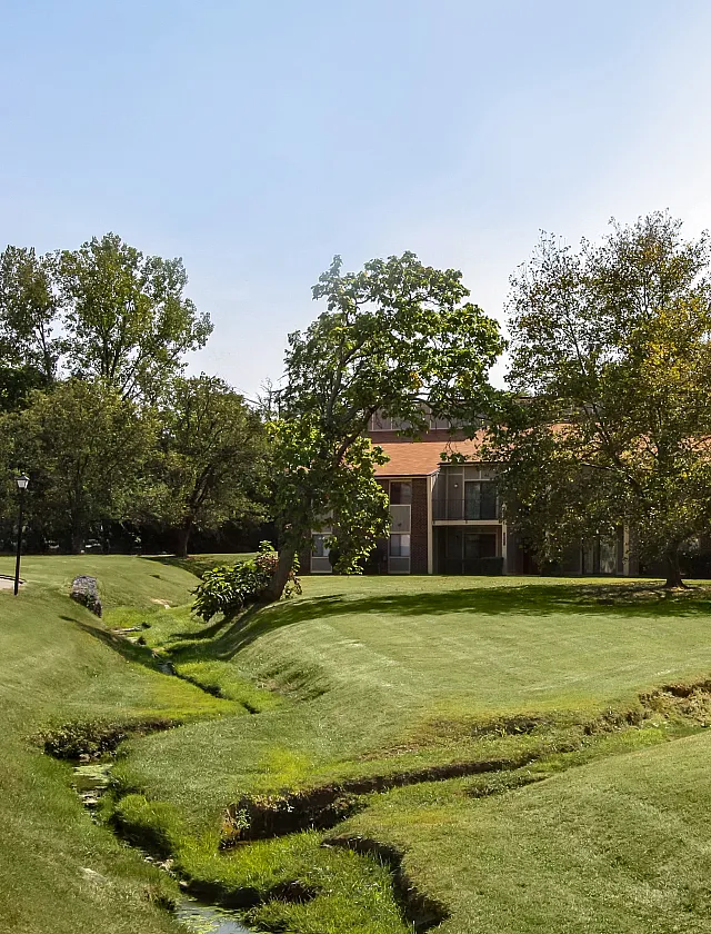 Charming apartment with scenic landscaped lawn and mature trees, featuring a serene creek under a clear blue sky.