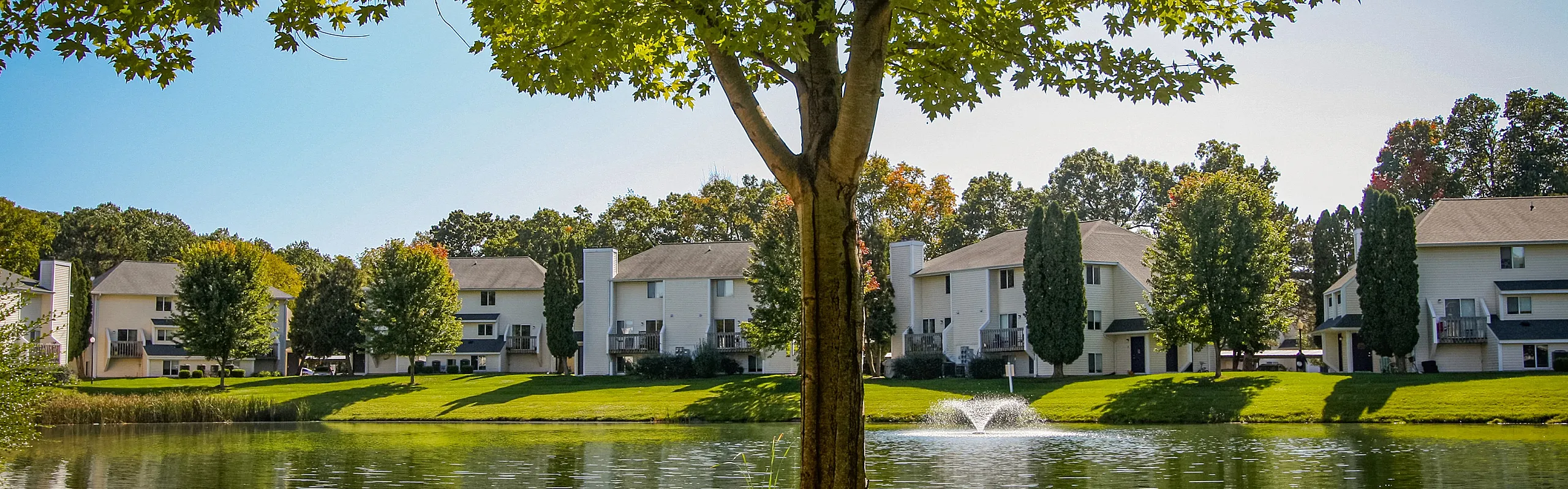 Charming lakeside apartment complex surrounded by lush greenery and vibrant trees, offering serene water views with fountain.