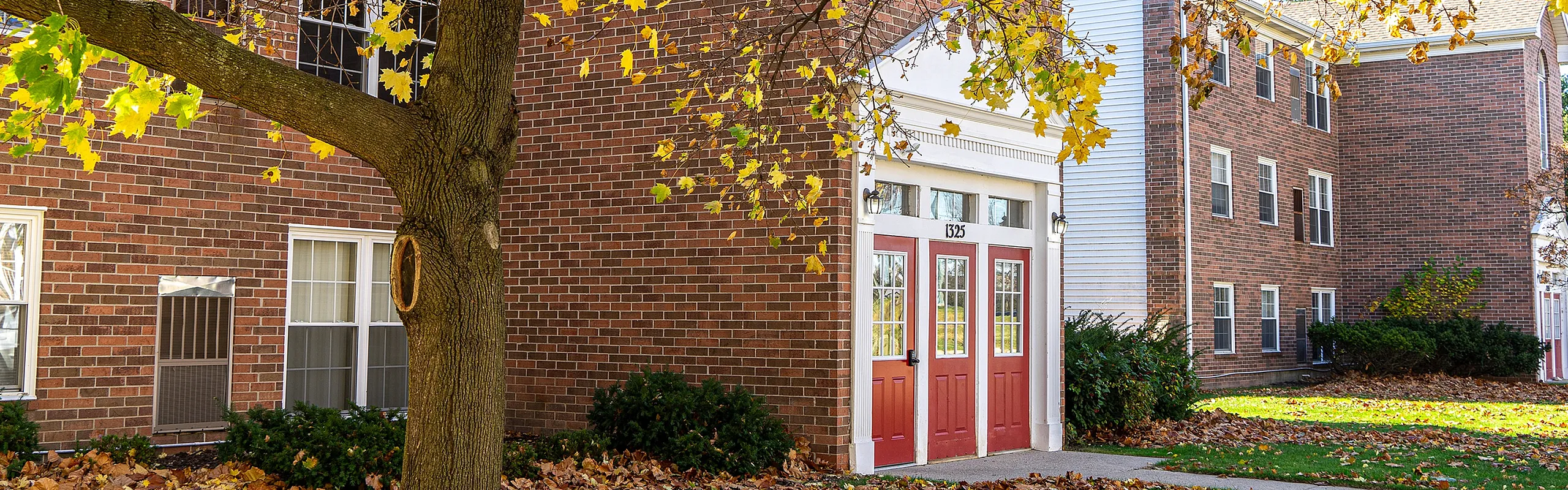 Charming brick apartment building with red doors, surrounded by lush autumn foliage and a picturesque landscape.