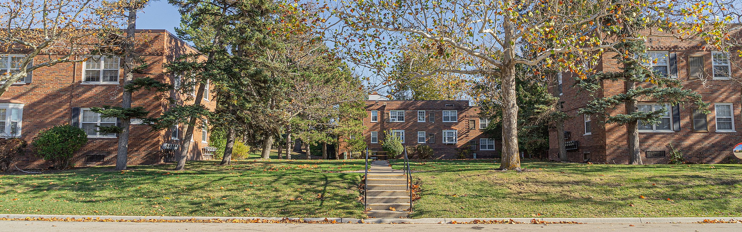 Charming brick apartment buildings surrounded by lush green lawns and mature trees beneath a clear blue sky.