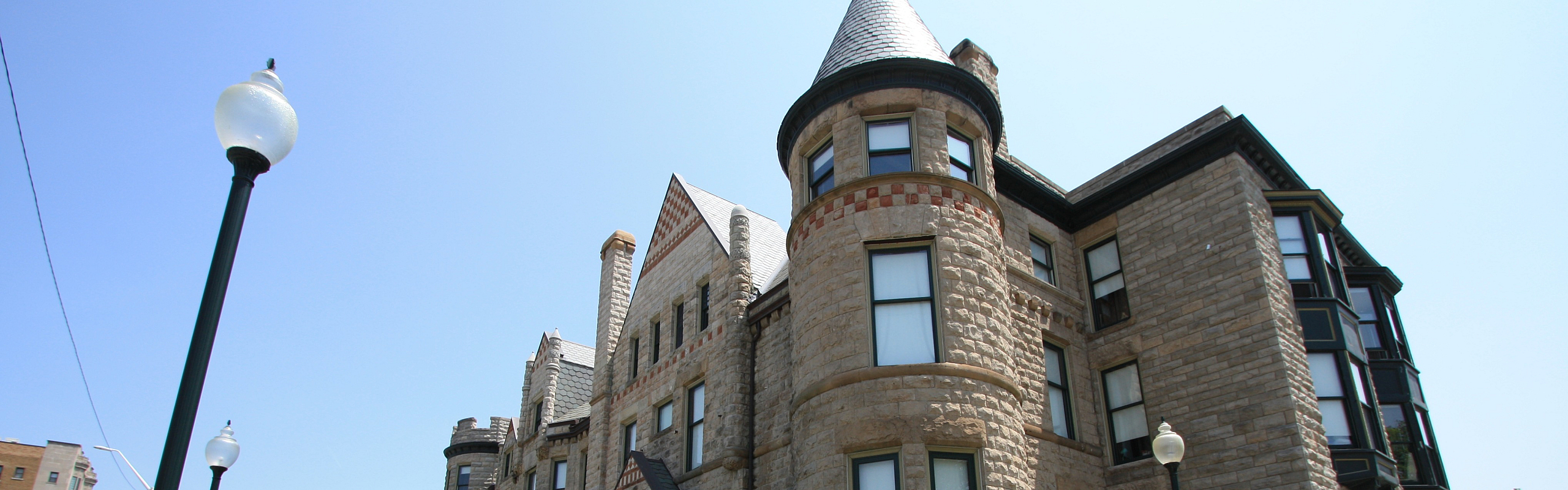 Charming historic building with a castle-like facade, featuring stonework and elegant turrets, surrounded by manicured landscaping and a wrought iron fence.