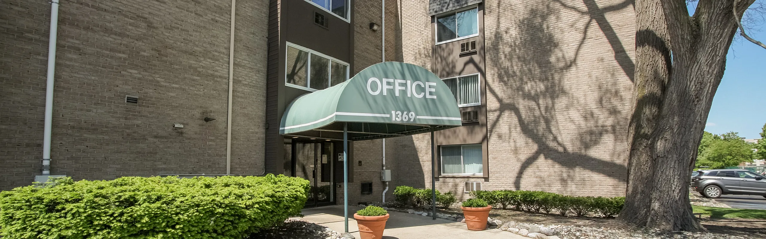 Welcoming apartment building entrance with lush greenery, charming brick exterior, and shaded walkway under a stylish awning.