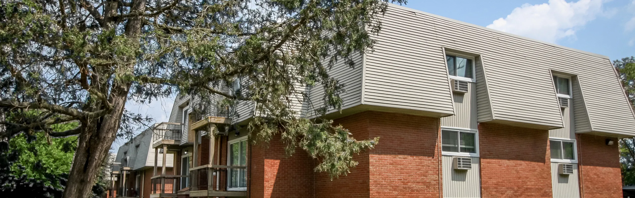 Charming multi-unit brick apartment building with unique sloped roofs, nestled in a lush green setting under a clear blue sky.