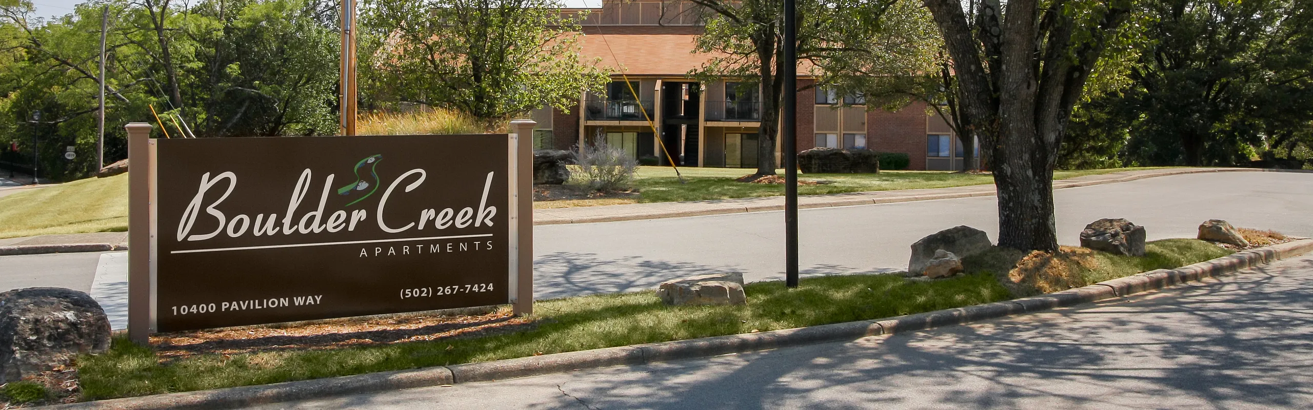 Charming Boulder Creek Apartments entrance surrounded by lush greenery and elegant trees, inviting peaceful living.