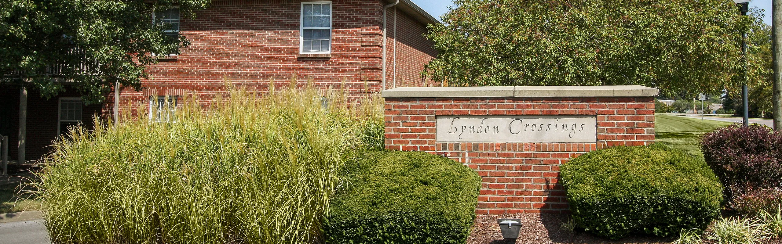 Inviting entrance to Lyndon Crossings with lush greenery and classic brick architecture, offering a serene suburban setting.