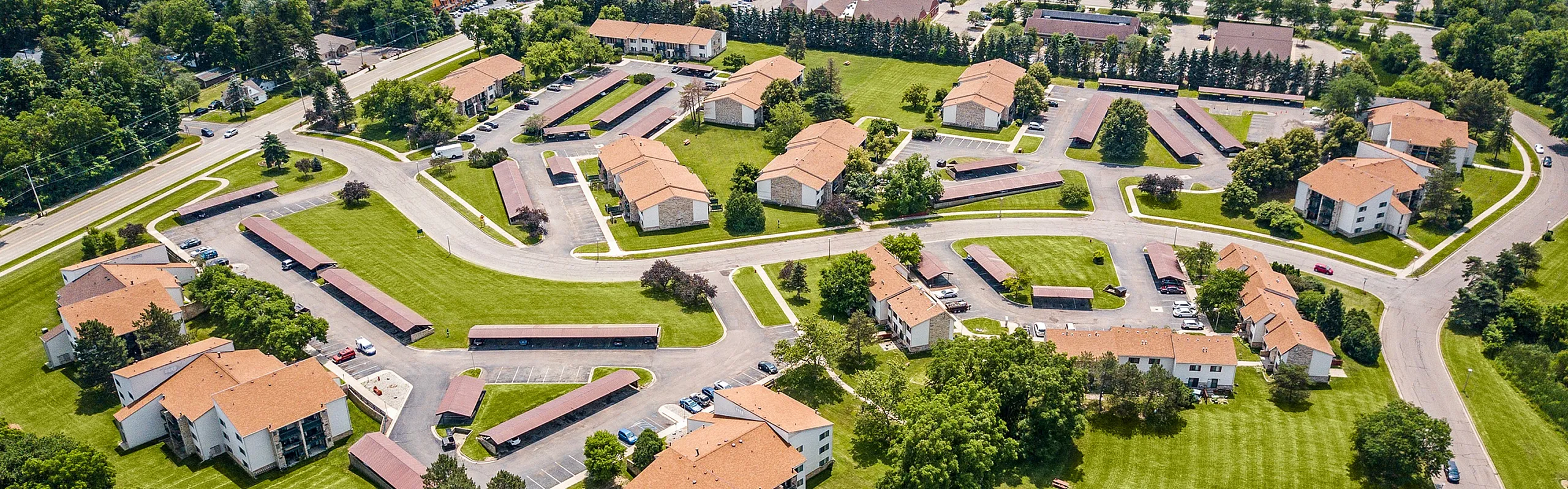 Aerial view of a picturesque apartment complex with beautifully landscaped grounds, charming red-roofed buildings, ample parking, and a pristine swimming pool.