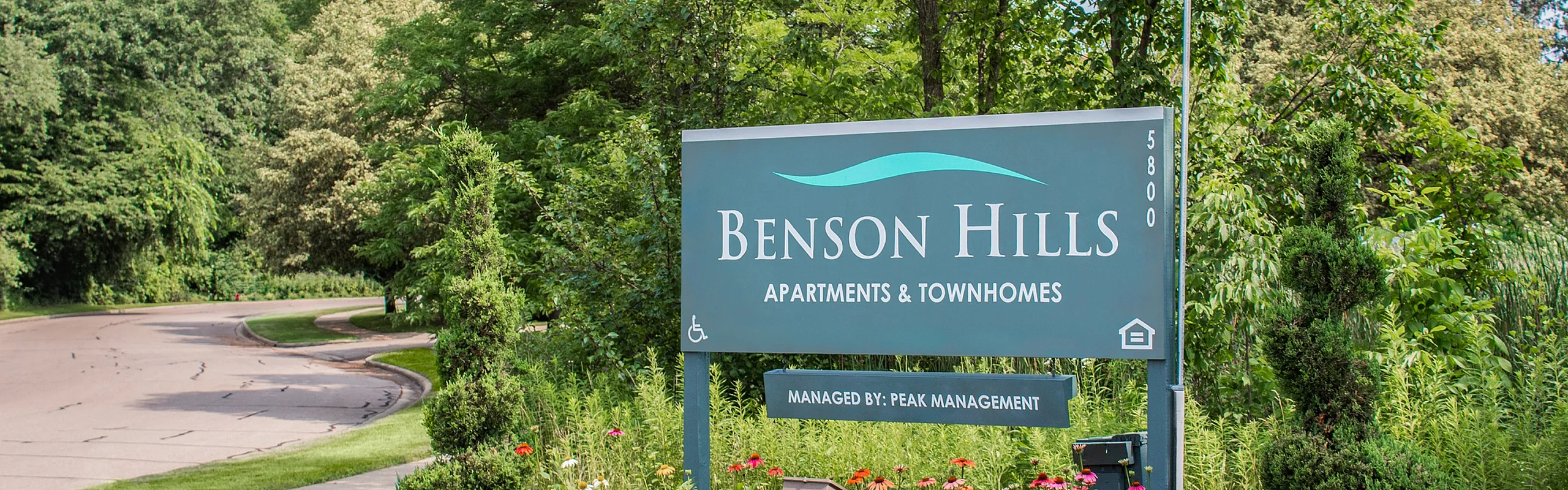 Inviting entrance to Benson Hills Apartments & Townhomes surrounded by lush greenery and vibrant flowers.