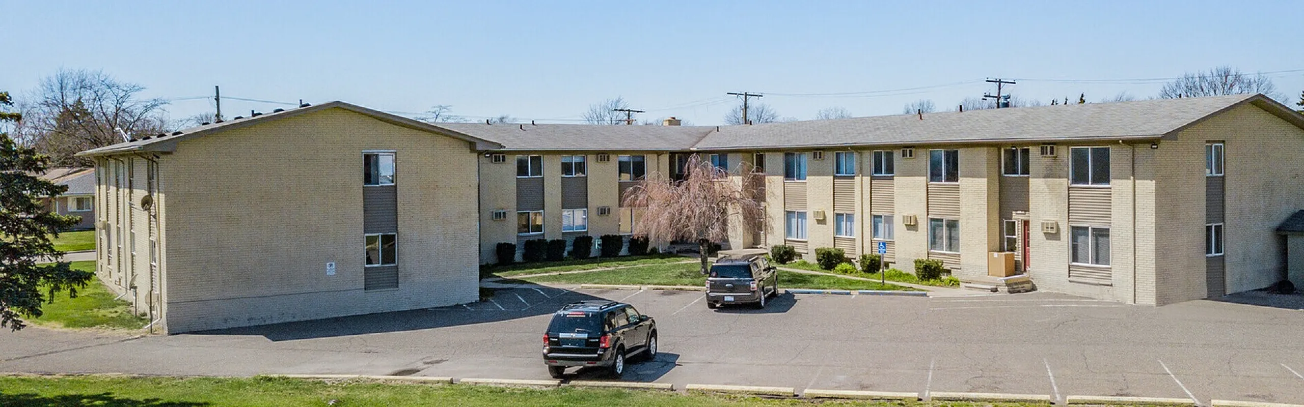 Beautiful two-story apartment building with ample parking, surrounded by lush green landscape and clear blue skies.